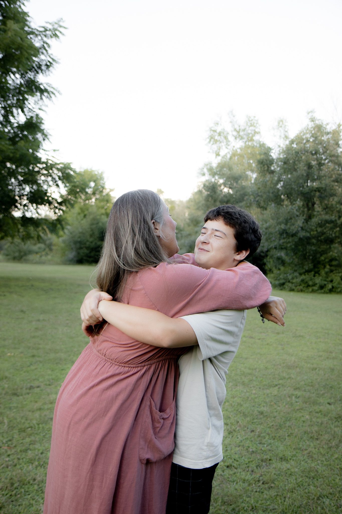 Two people share a warm hug outdoors in a green park, one smiling joyfully, surrounded by lush trees.