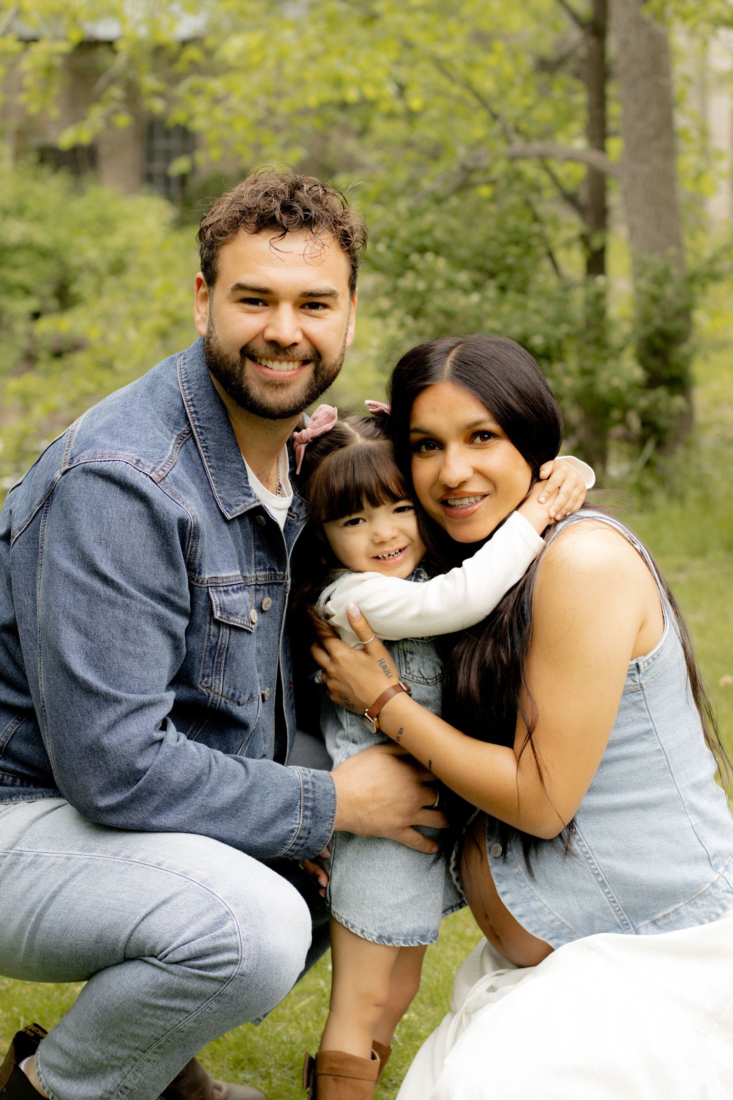 A smiling family of three poses outdoors, with parents hugging their young daughter in a lush green park setting.