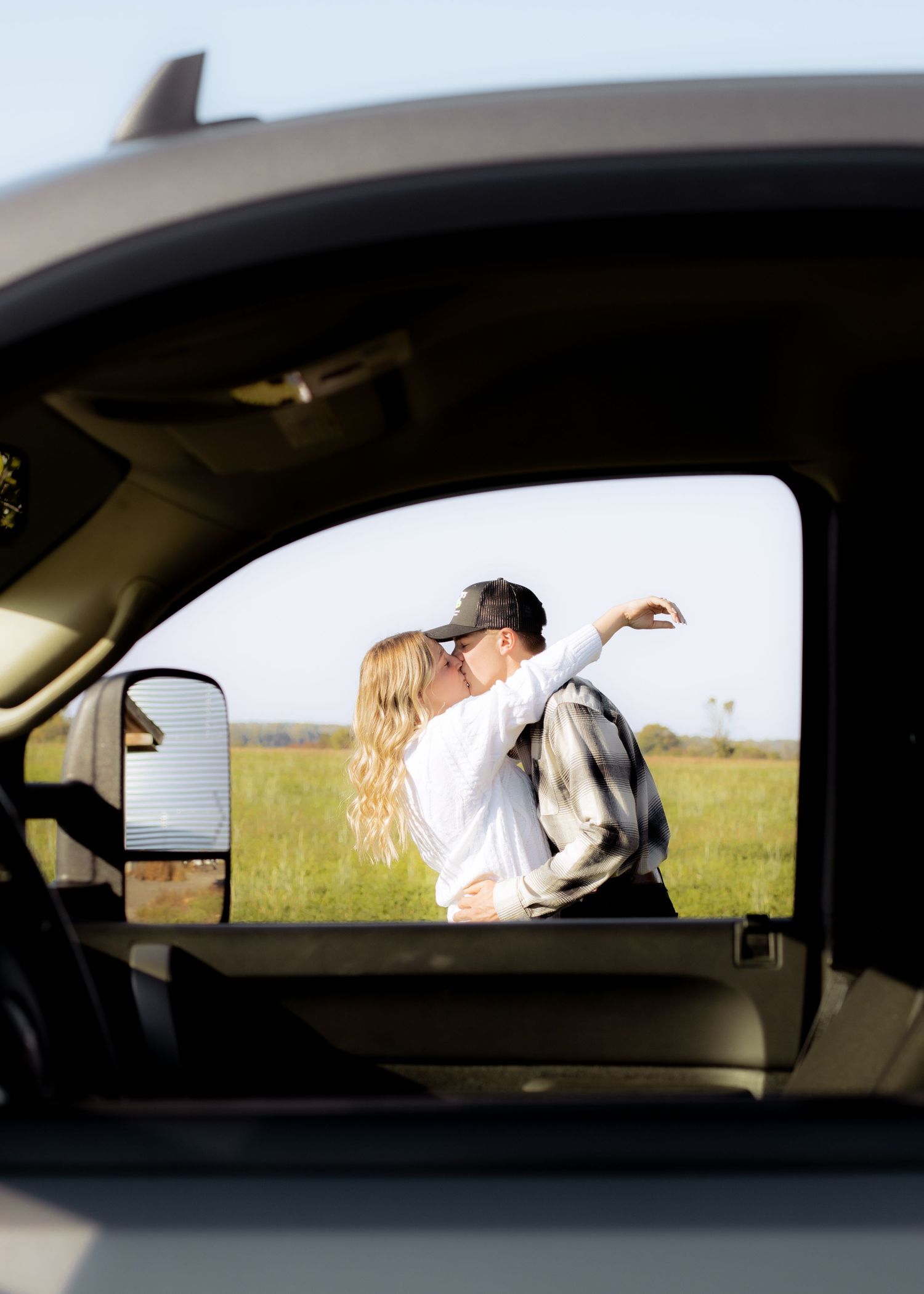 Couple sharing a kiss in a green field captured through the window of a pickup truck.