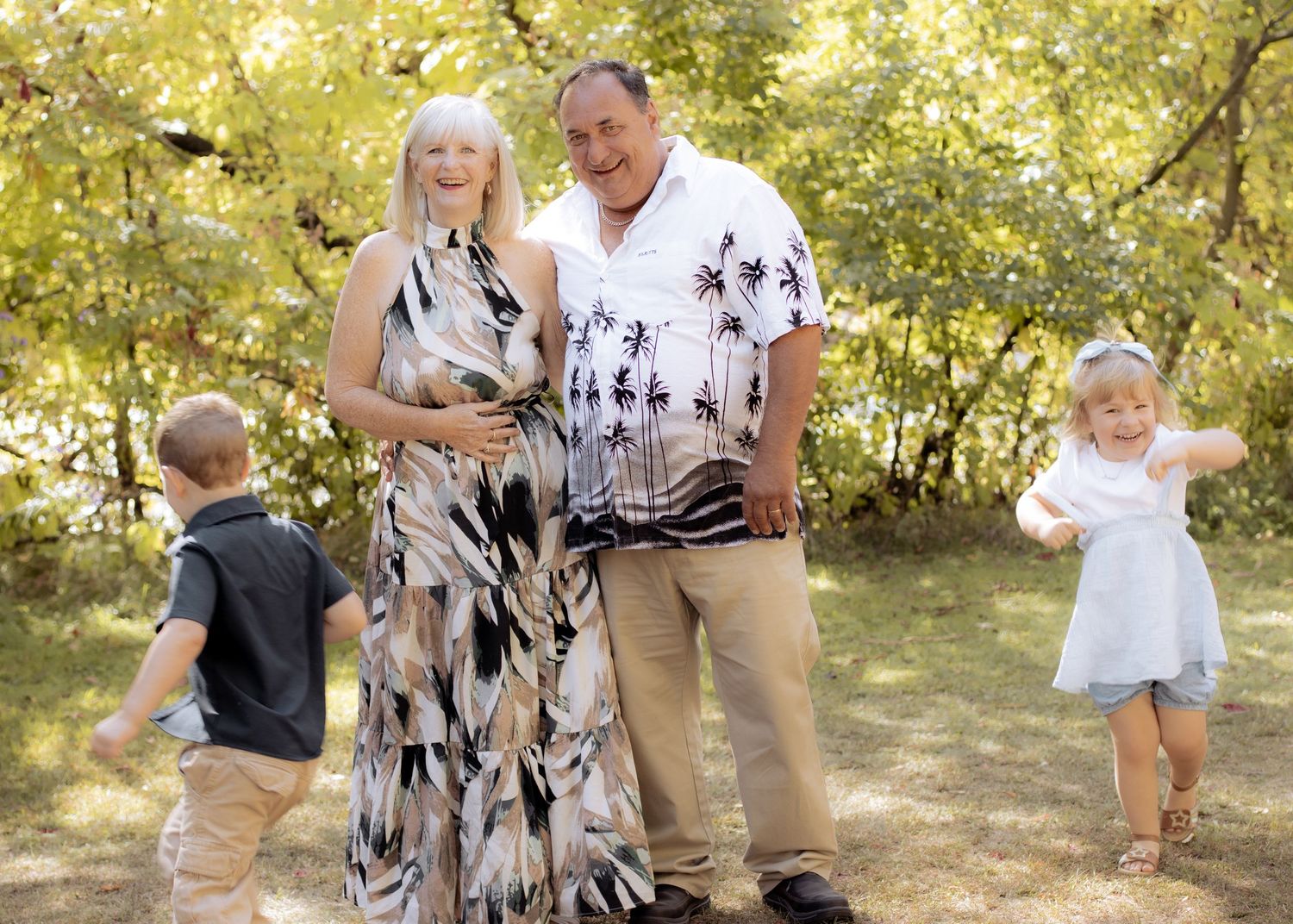 Grandparents laughing outdoors as two young grandchildren play around them in golden park setting.