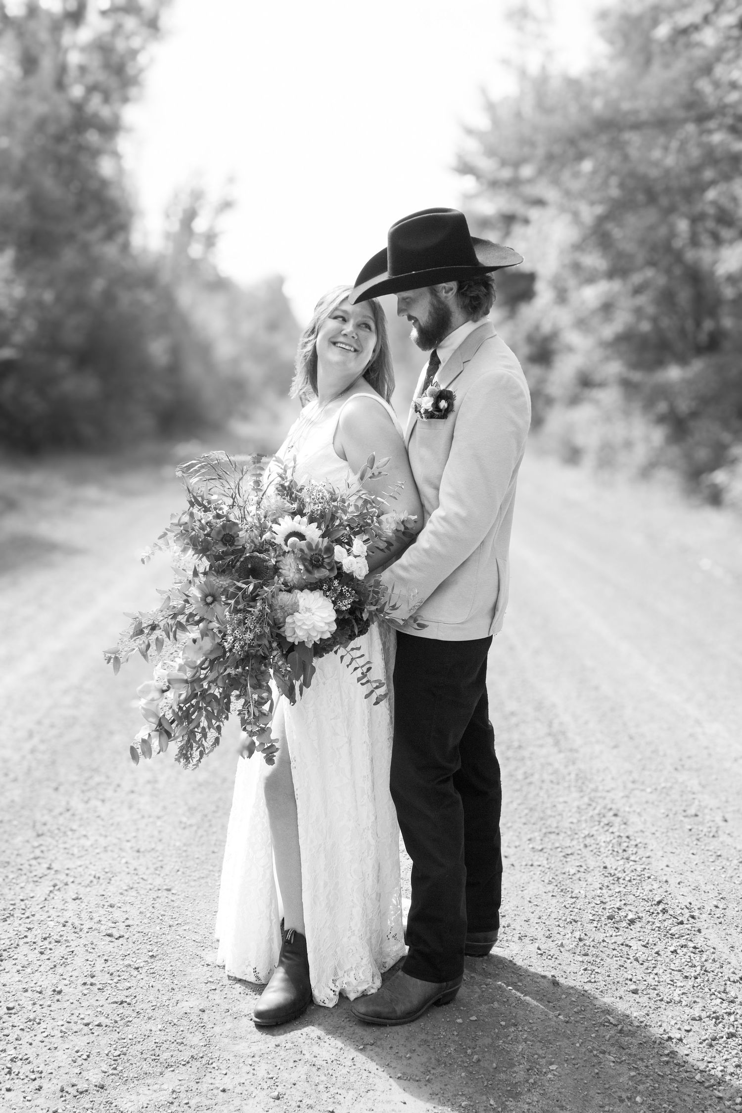 A black and white photo of a bride and groom on a dirt road, groom wearing a cowboy hat.