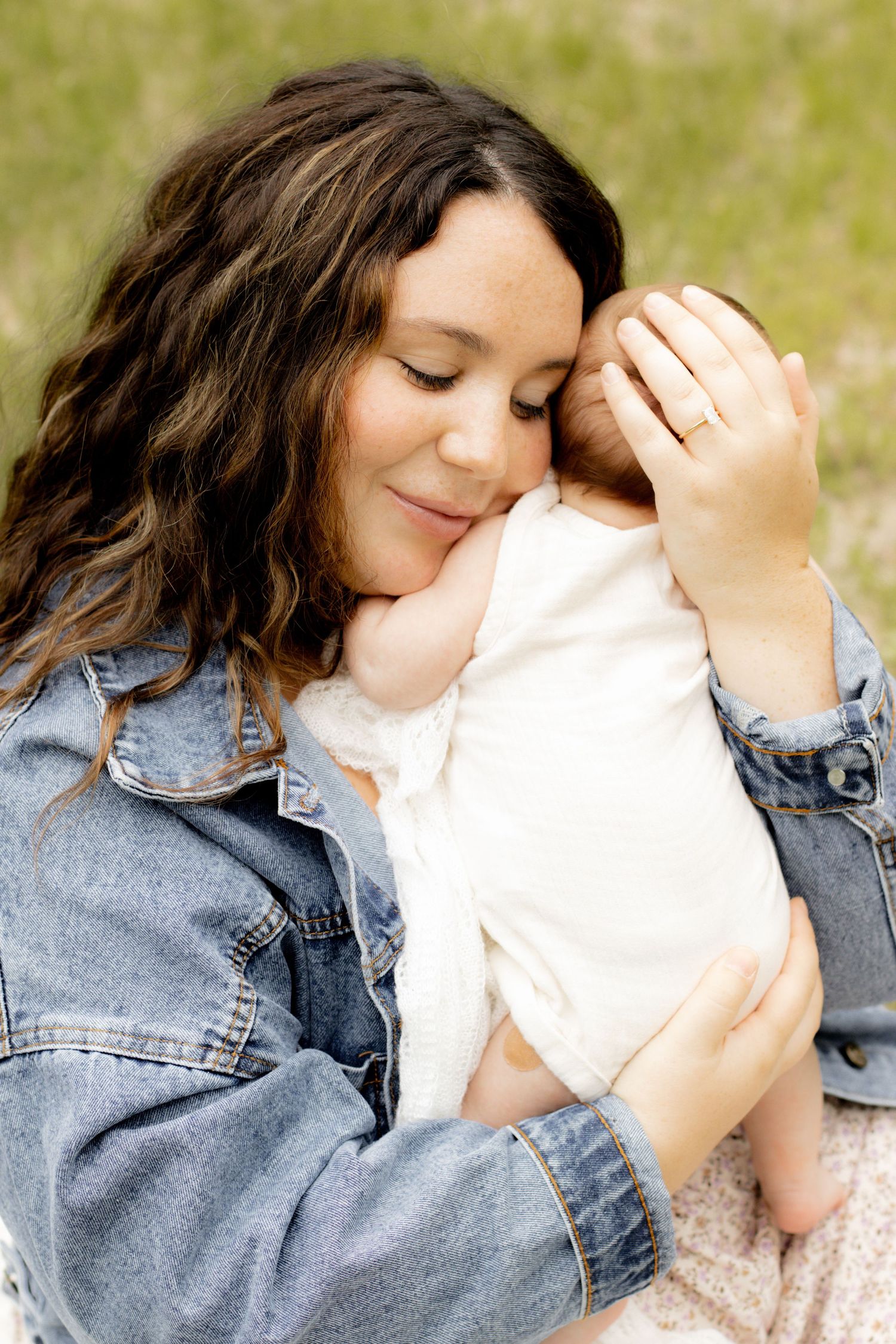 A mother with wavy brown hair tenderly holds her newborn baby close to her face outdoors.