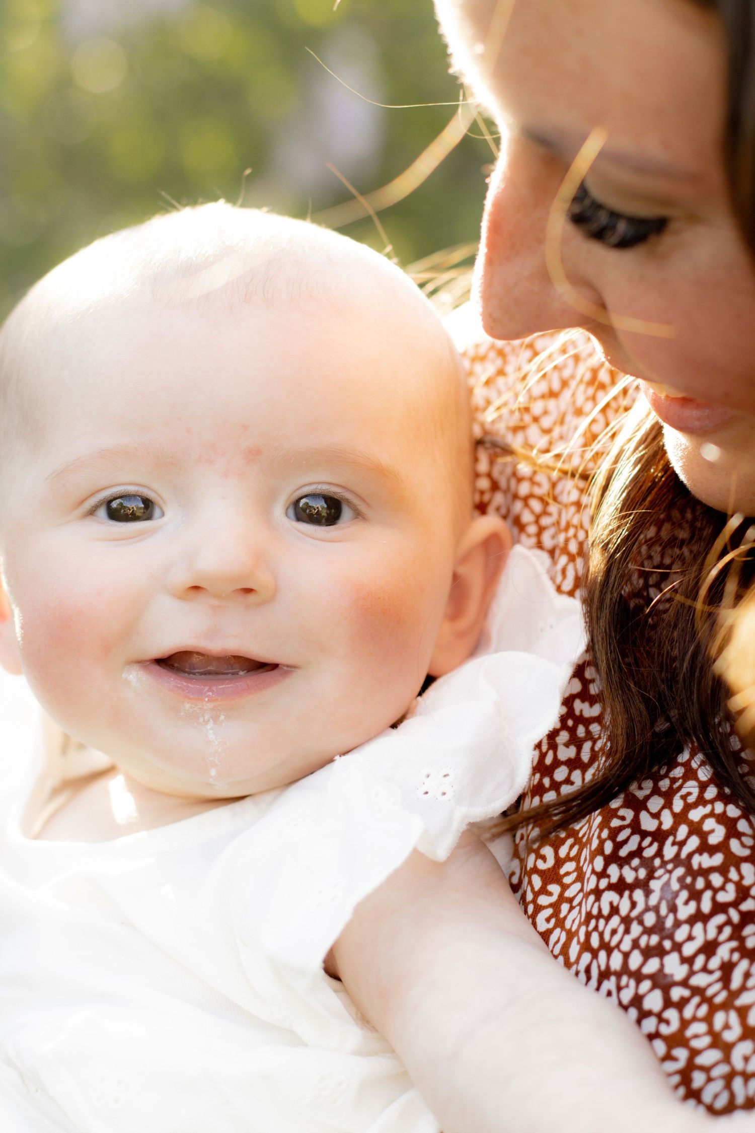 A mother gazes lovingly at her smiling, bright-eyed baby in warm sunlit outdoor portrait.