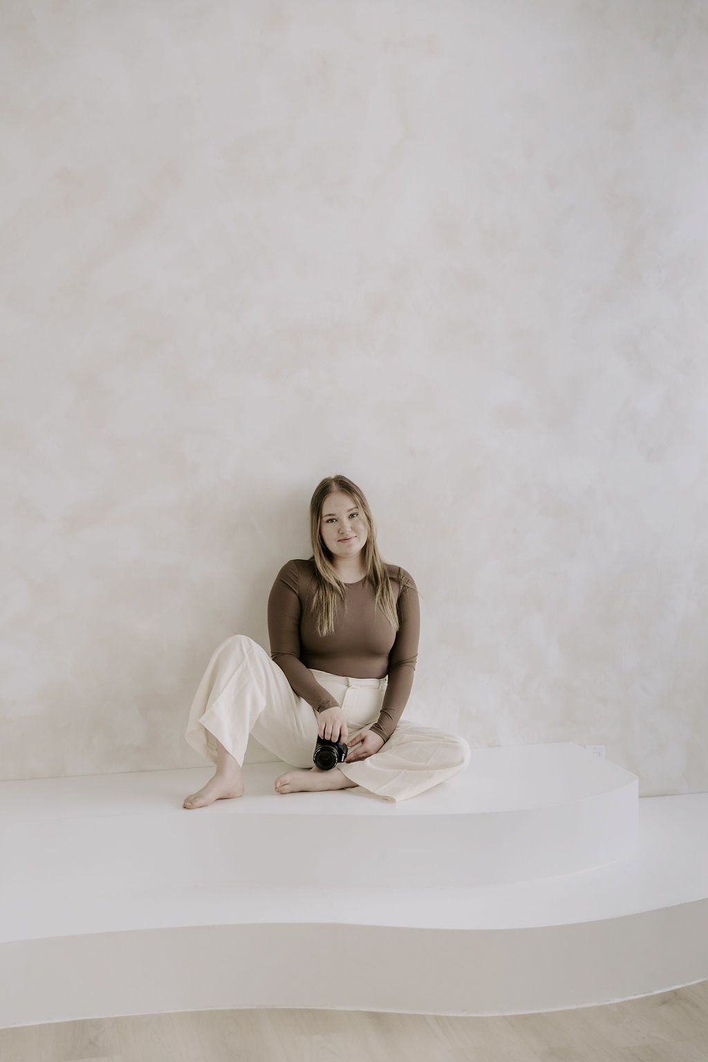 Young woman in brown top and white pants sitting cross-legged holding camera in white studio.