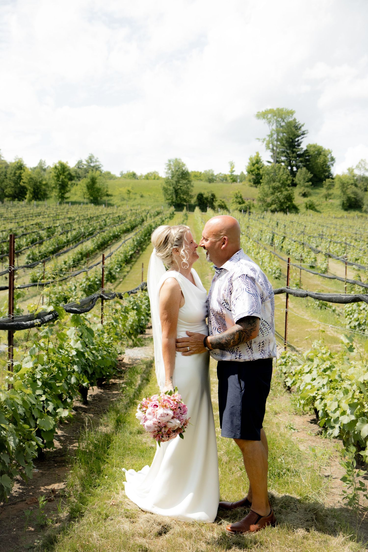 Bride and groom share a romantic moment in a lush vineyard, bride holding a pink bouquet in a white gown.