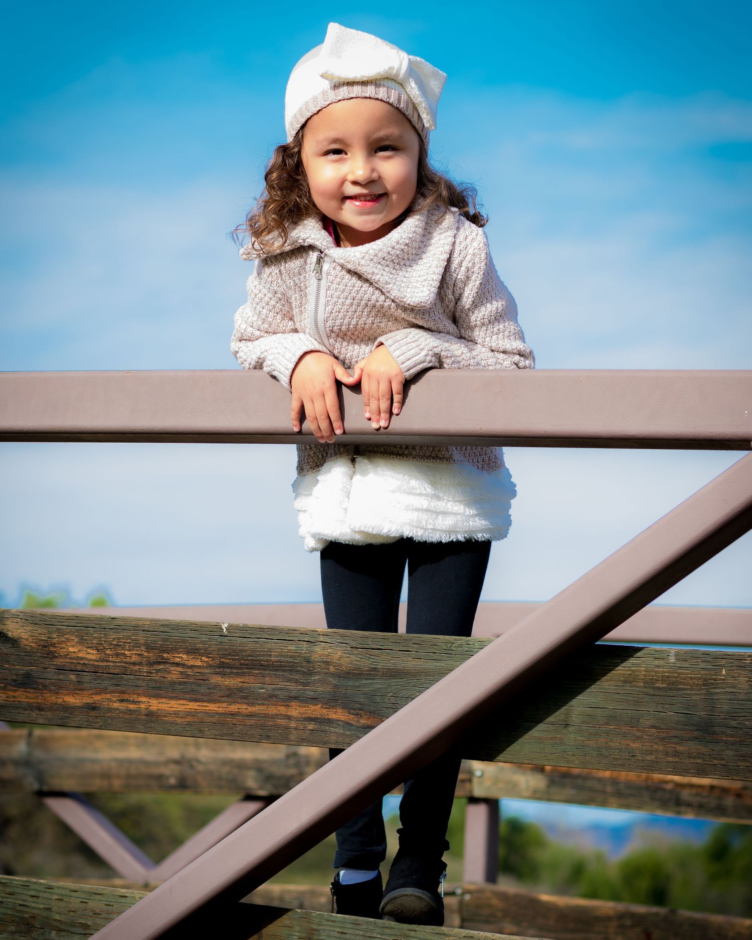 Child in white sweater looks over wooden railing with blue sky and landscape in background.