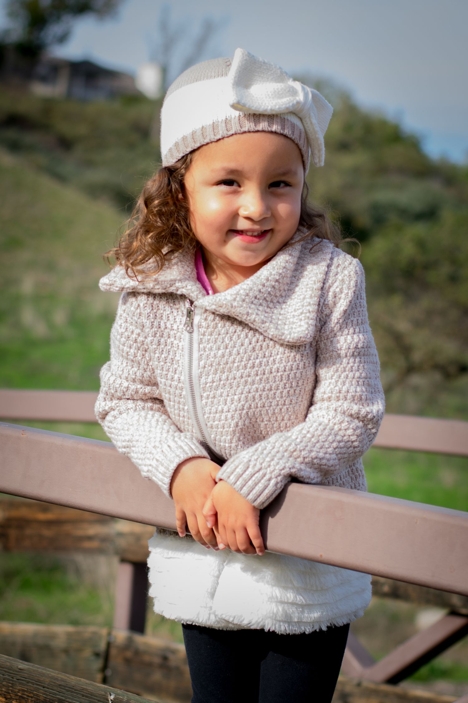 Small child in white knit sweater and cap smiles while leaning on wooden railing outdoors.