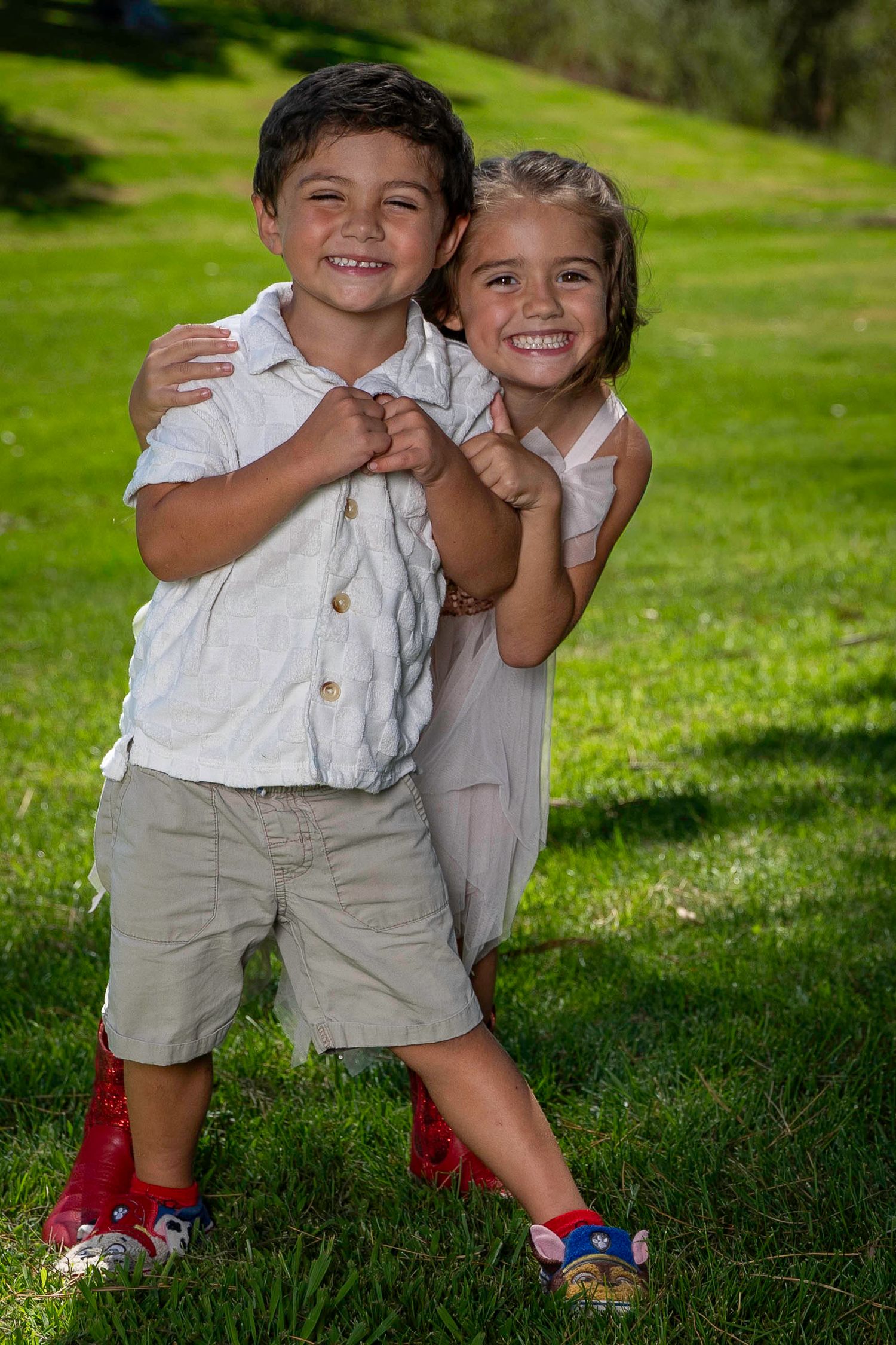 Two children in white outfits share a playful moment while standing on green grass during a sunny day outdoors.