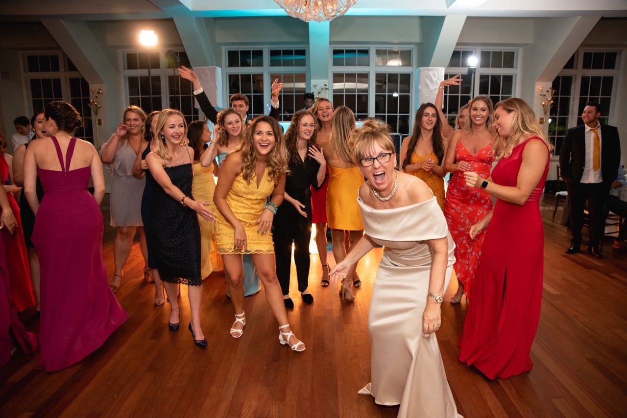 Joyful wedding reception guests dancing together on a wooden dance floor under blue ambient lighting.