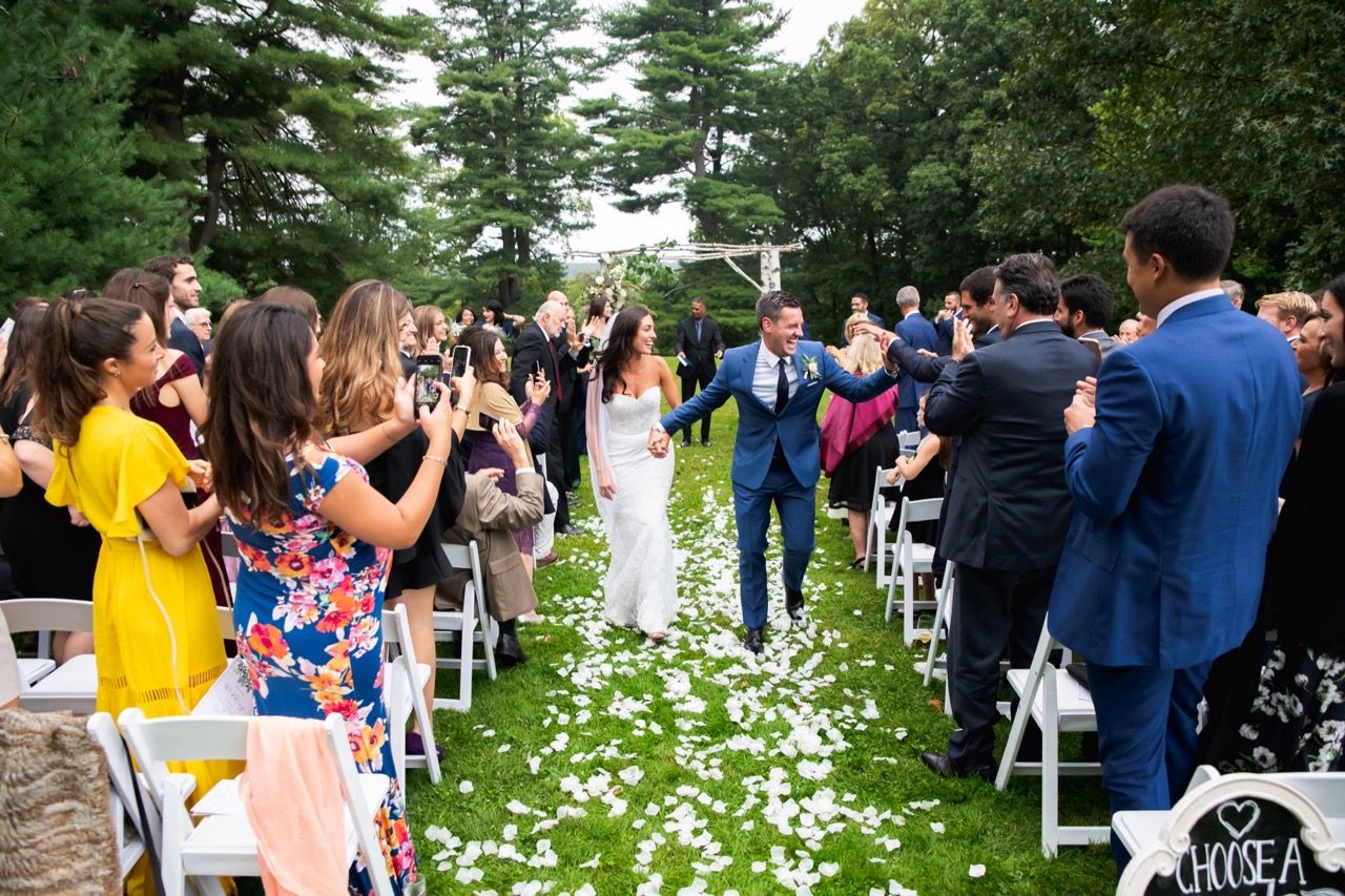 Outdoor wedding ceremony with white flower petals lining the aisle under green trees.