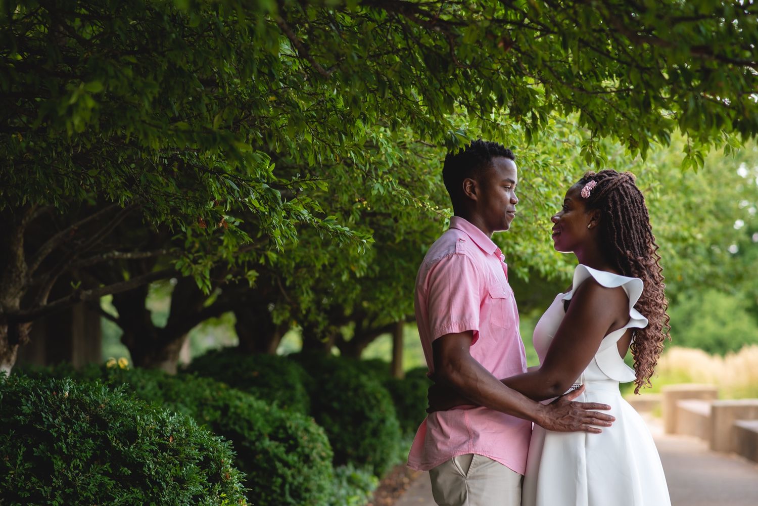 Couple walking together under lush green trees in a park during a summer engagement photography..