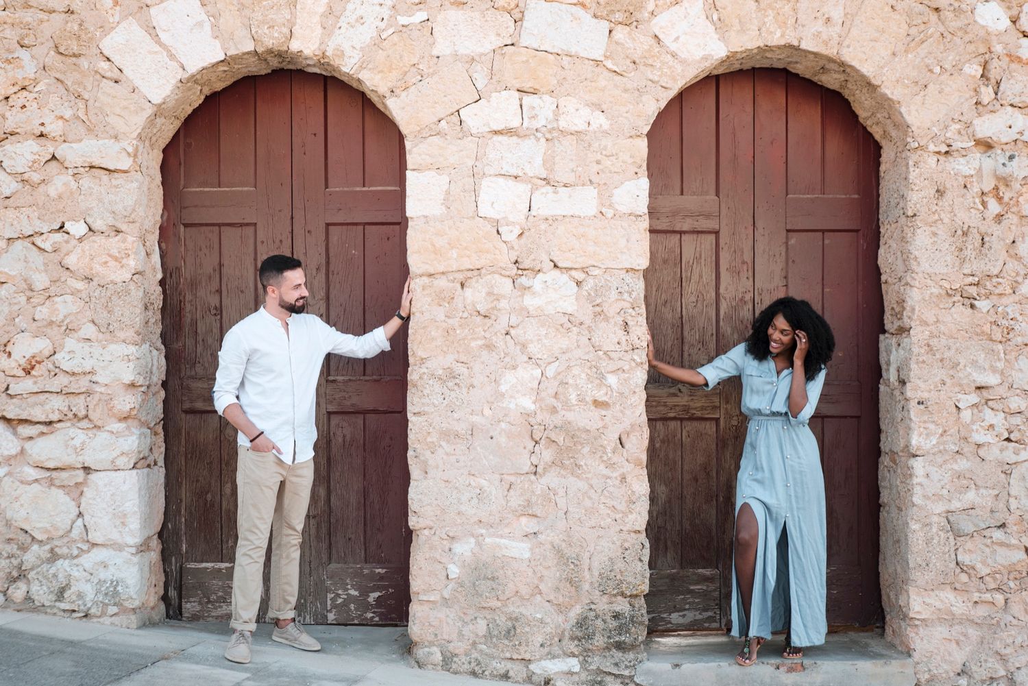 Playful couple posing at rustic stone archways with wooden doors during a couples engagement photography session in Spain.