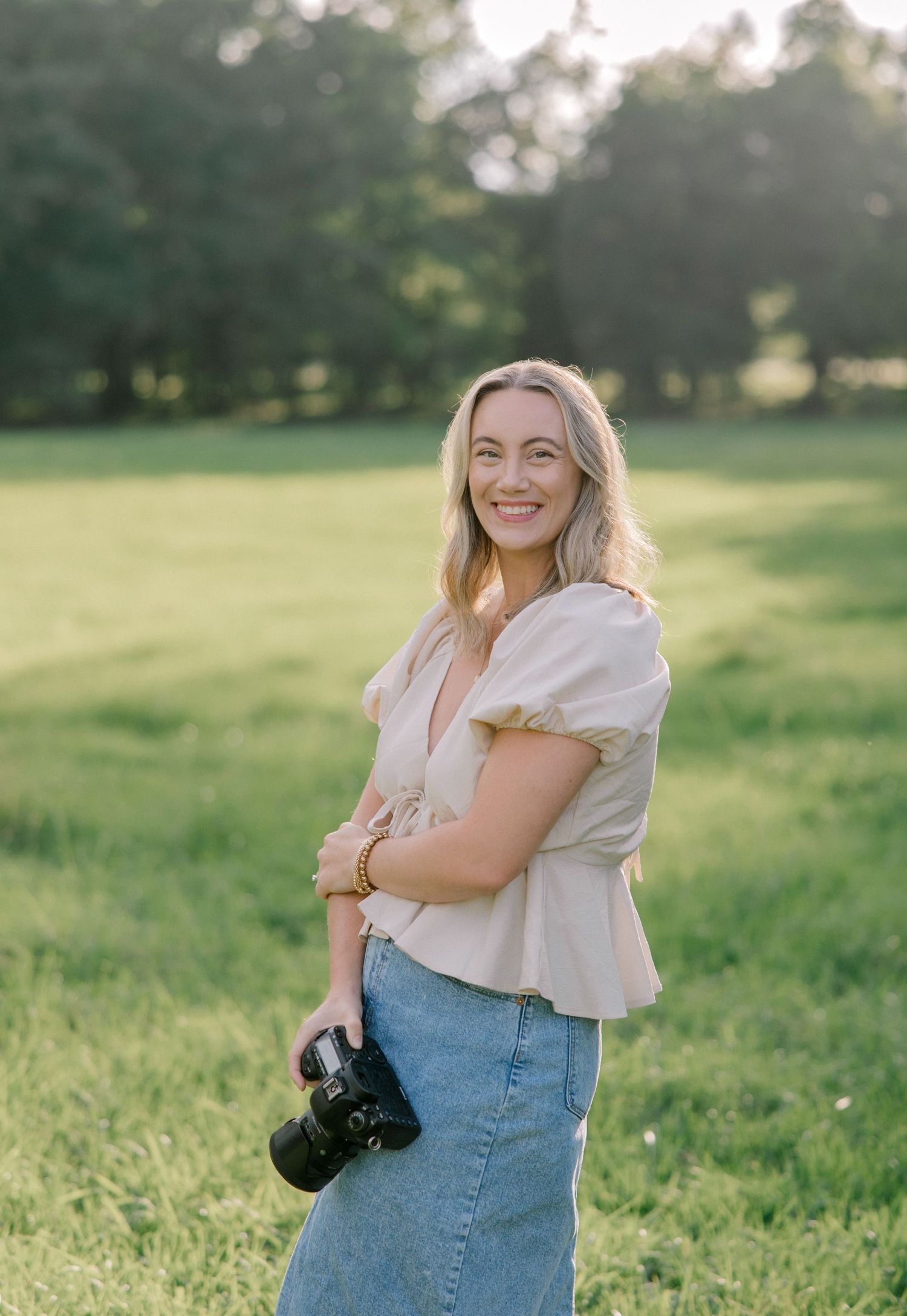 Photographer wearing blue denim skirt and white peplum top holds camera while standing in sunny green field.