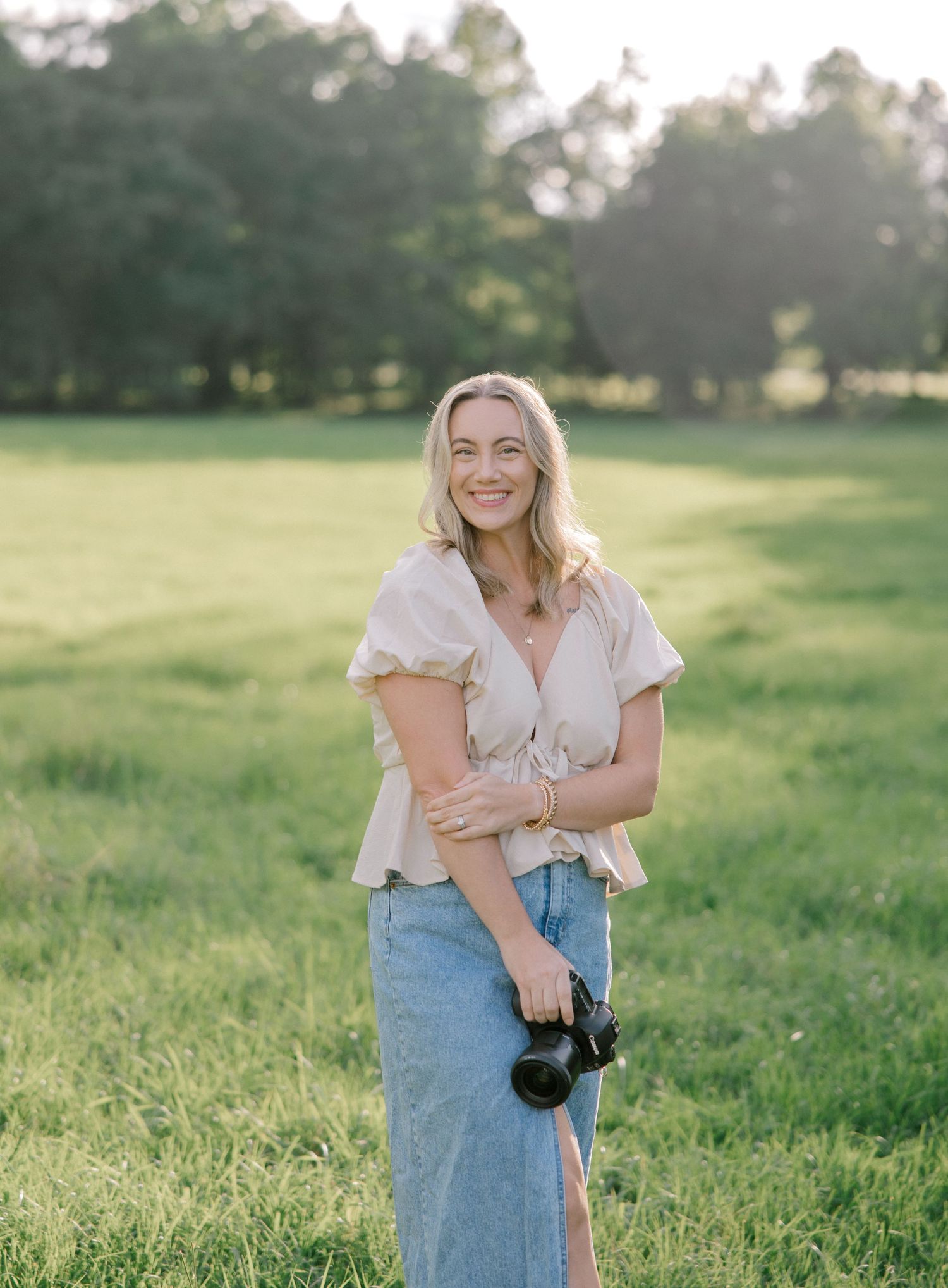 Smiling blonde woman holding a camera stands in a sunny green field wearing a beige top and denim skirt.