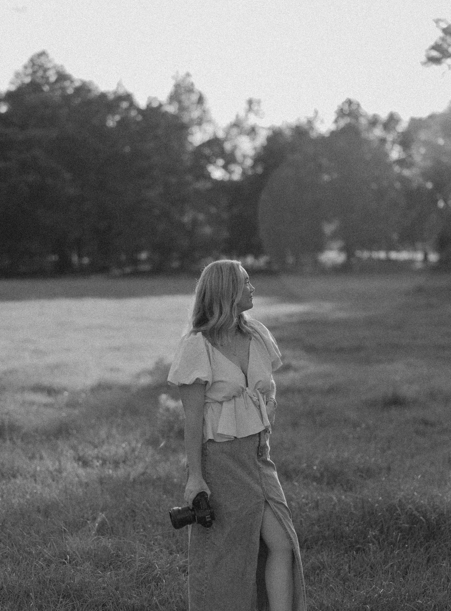 Black and white portrait of a person holding a camera in a flowing dress standing in a grassy field at dusk.