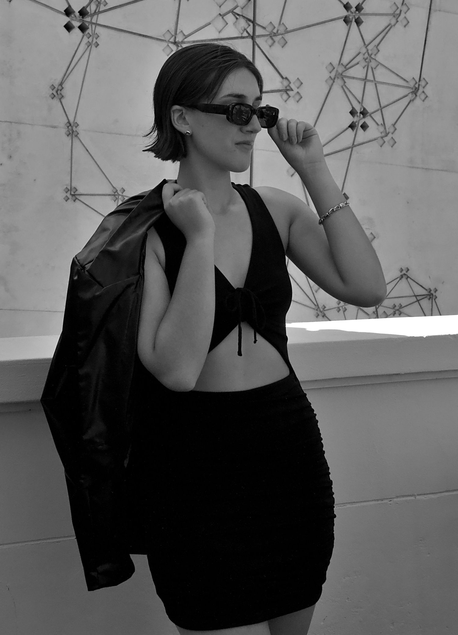 Fashionable woman leaning against a half wall, jacket over shoulder, wearing sunglasses, captured in a lifestyle content shoot at Wollongong