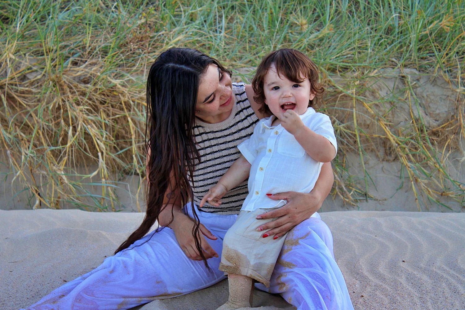 Beach motherhood session of a mother looking at her son as he smiles at the camera with grassy lands behind them
