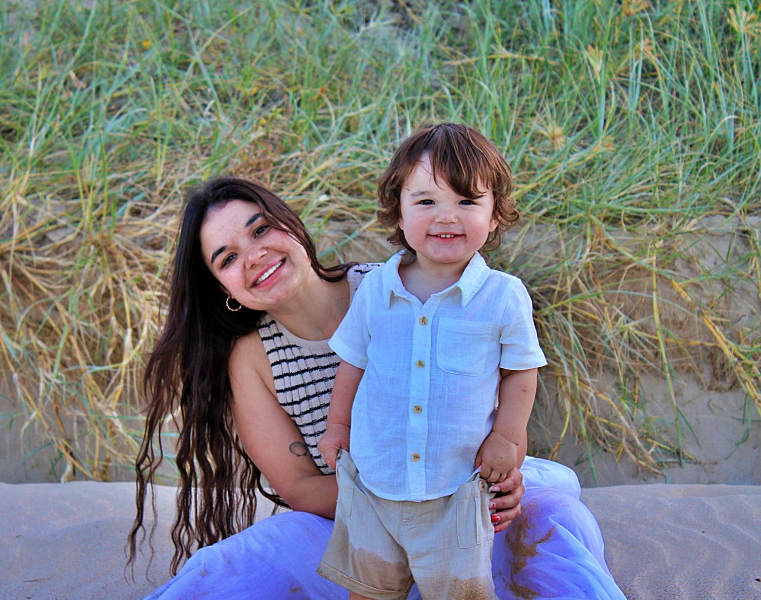 Mother and son smiling at the camera together while sitting on the beach during a motherhood session.
