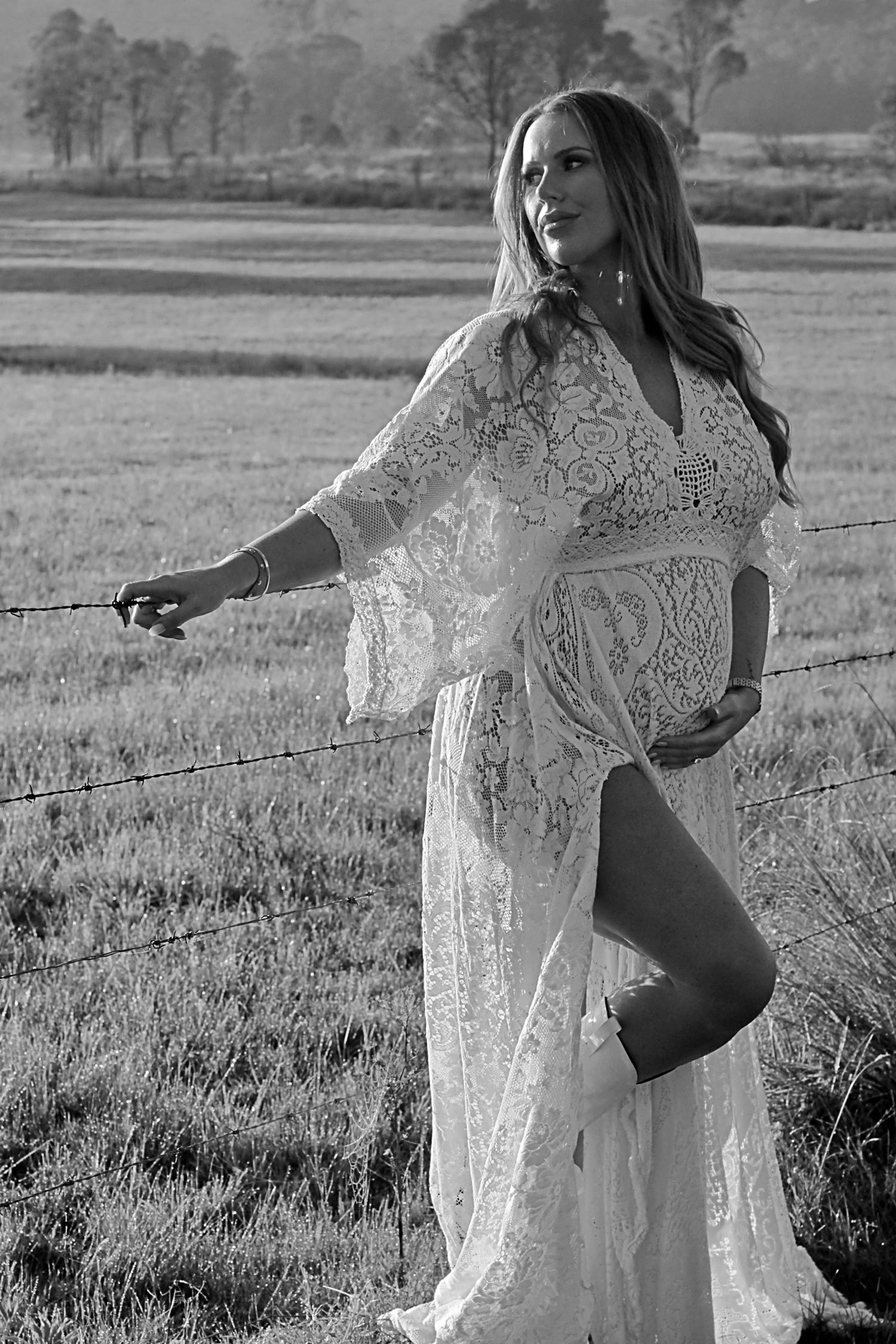 Black and white maternity portrait of a mother standing on a fence, one hand holding the fence, the other cradling her baby bump.