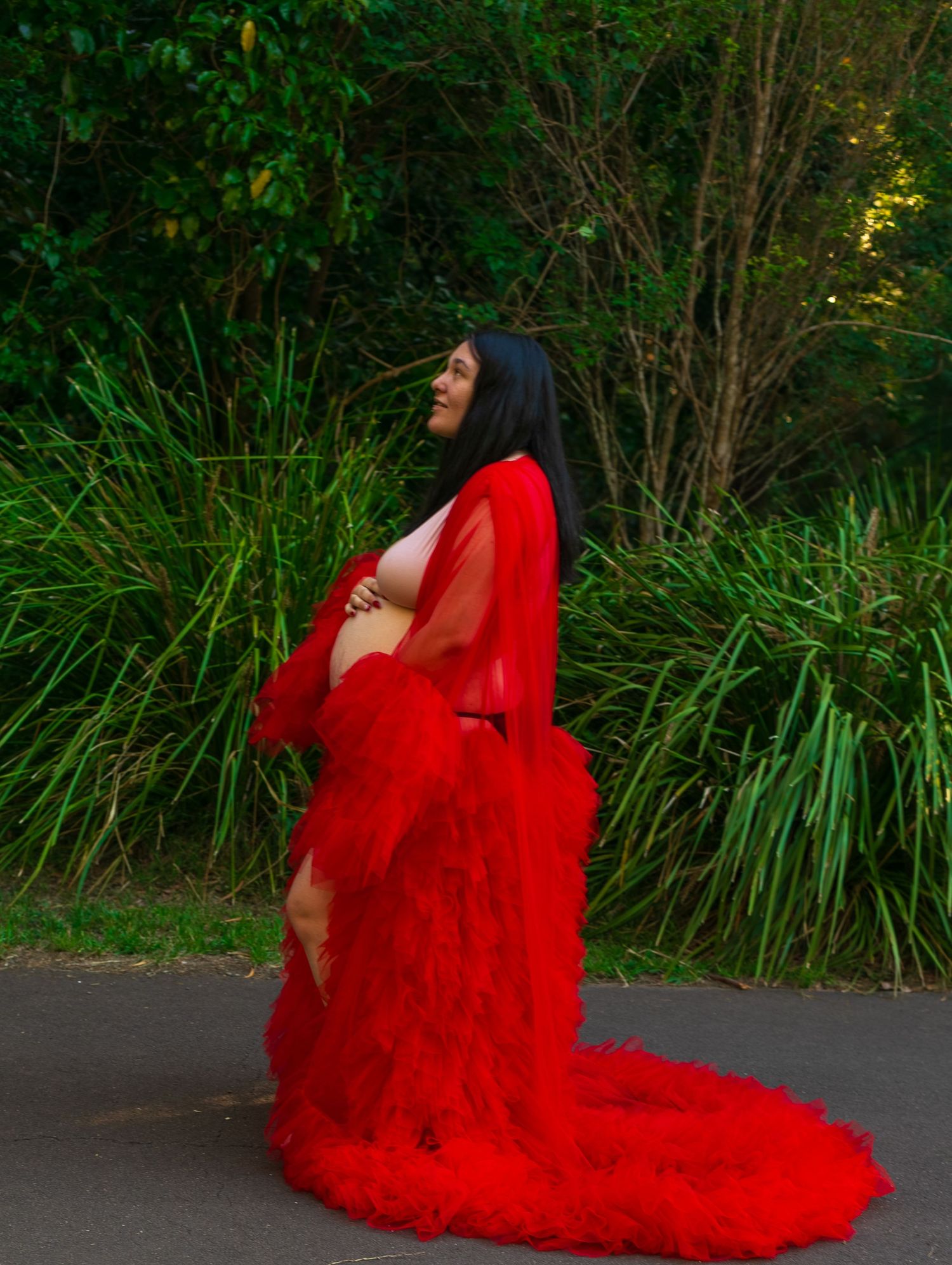 Mother-to-be in a red gown holding her bump, looking away from the camera with a forest background, capturing a serene moment.