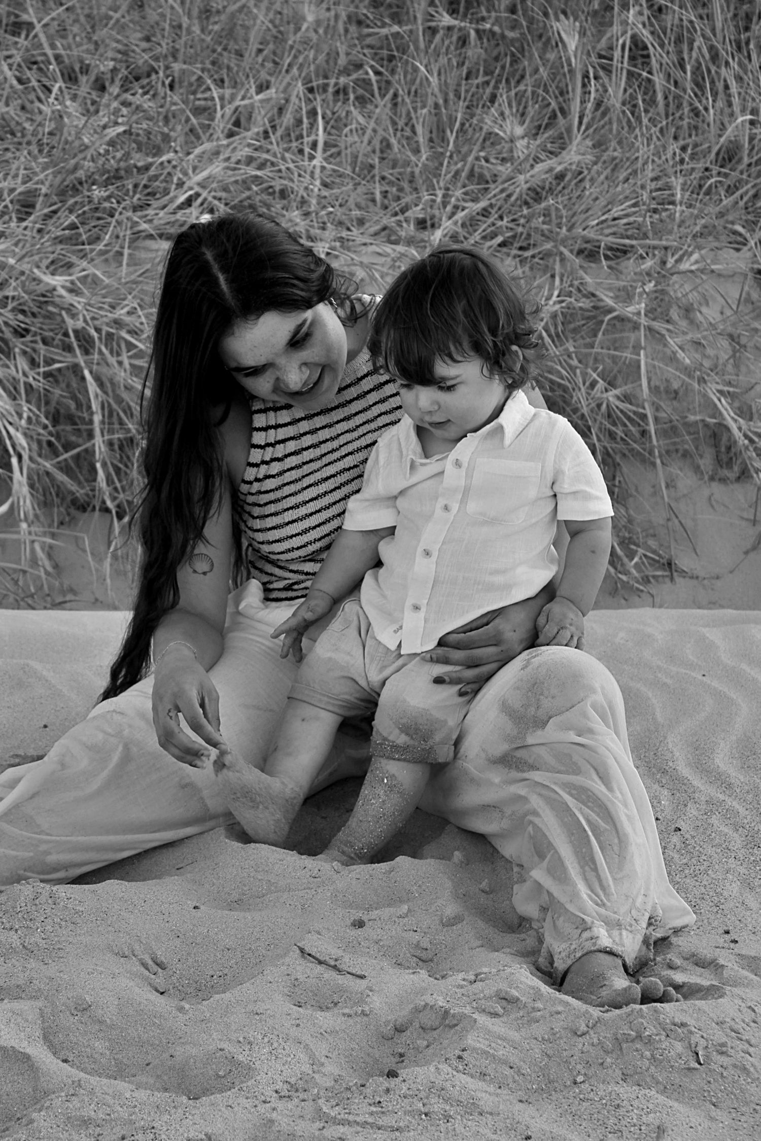 Motherhood session showing a mother and her son sitting on the beach with grassy lands in the background, playing with the sand together.
