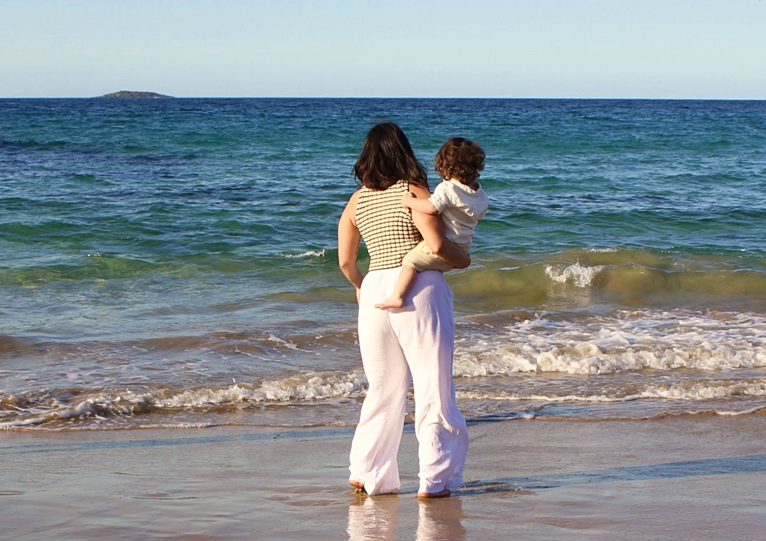 Mother holding her son on the shoreline during a beach motherhood session, looking out at the ocean, photographed from behind.