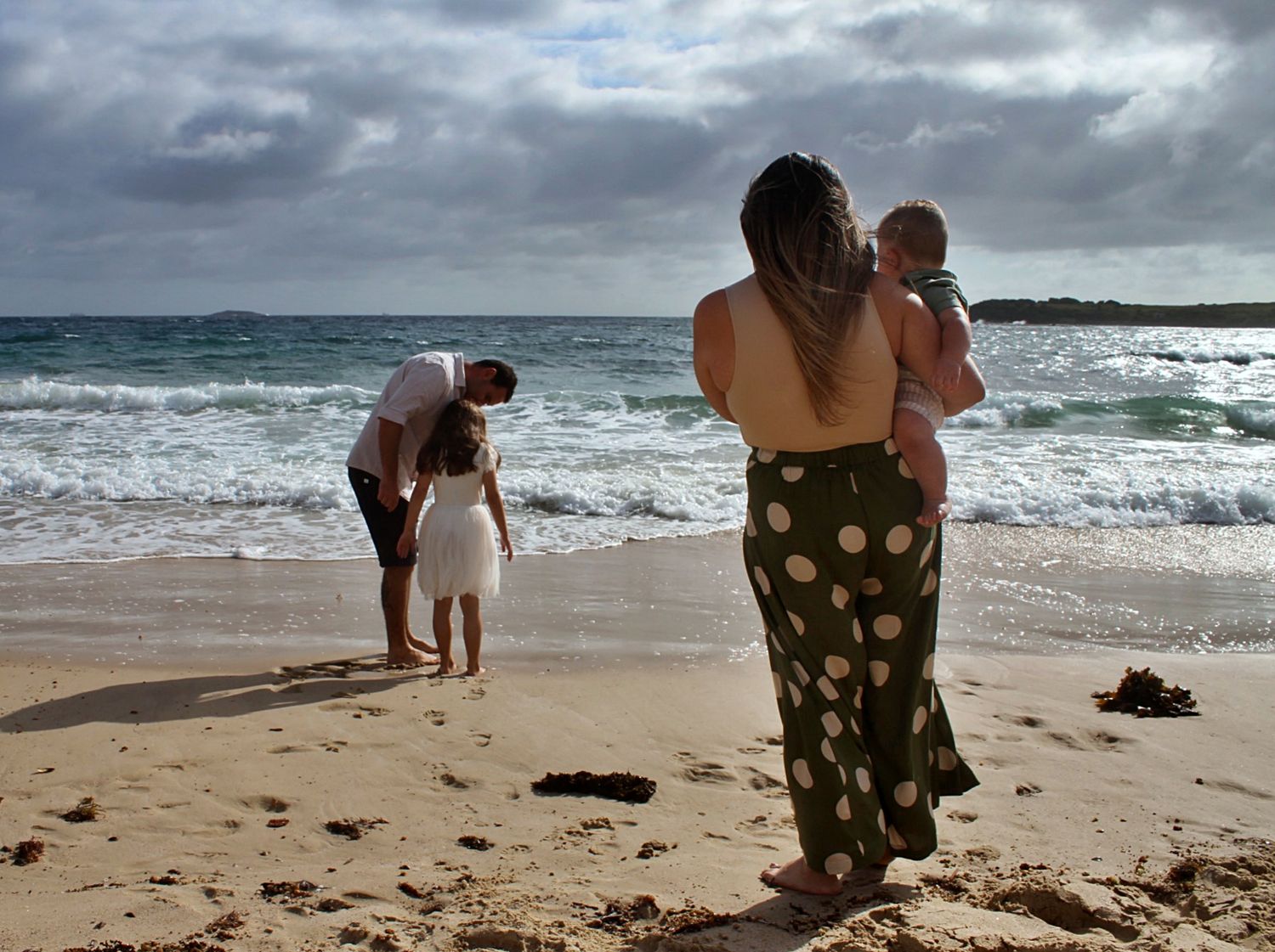 Mum and son watch dad and daughter play together on the beach in the Illawarra, capturing relaxed family connection by the sea.
