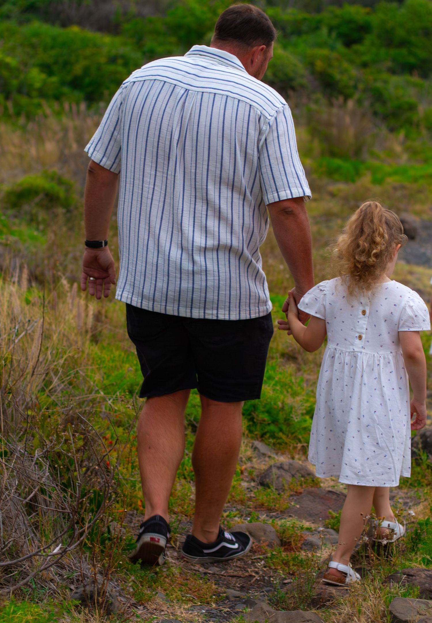 Dad holding his young daughter's hand as they walk together through a grassy coastal landscape.