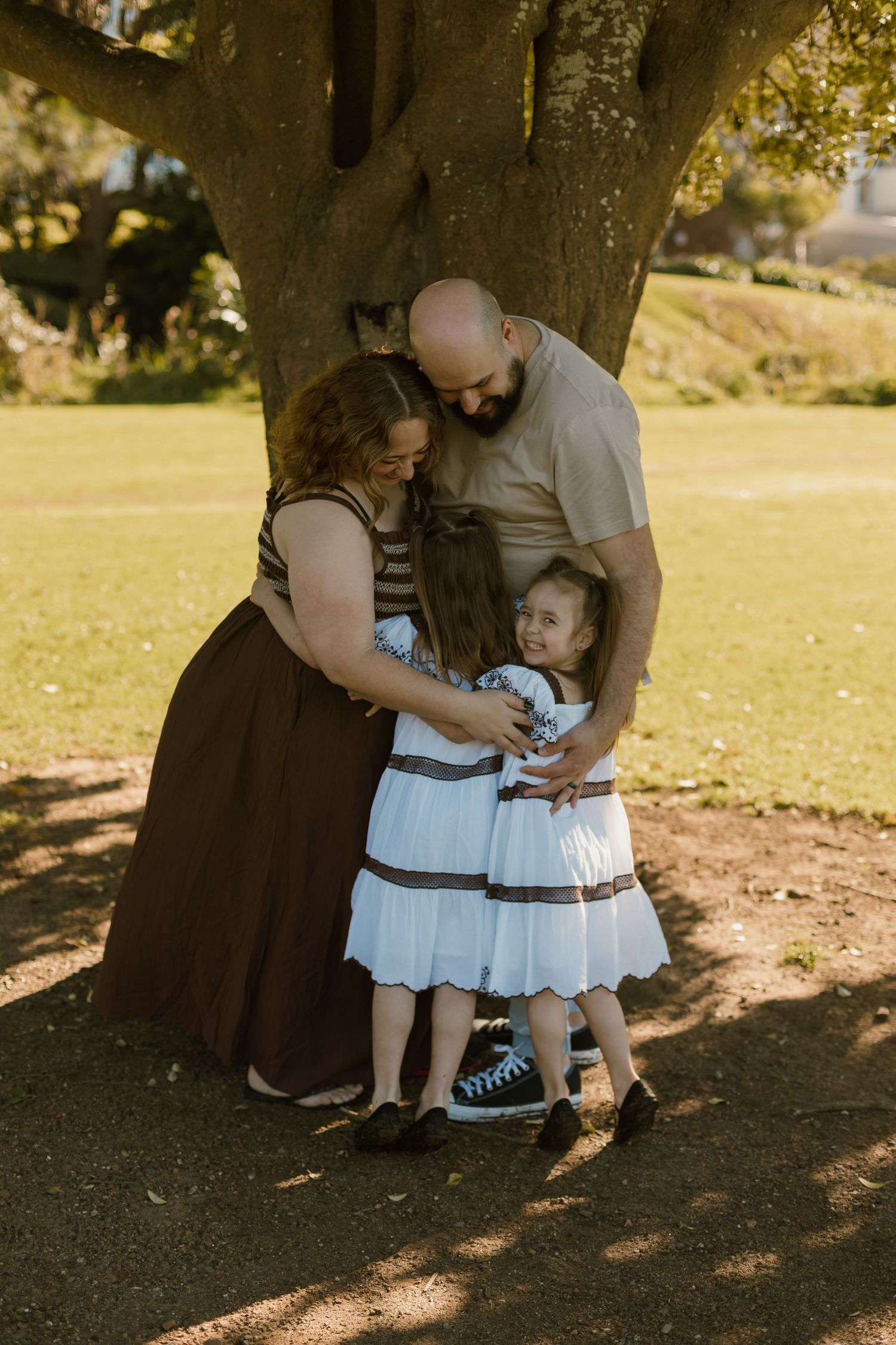 Mum and Dad cuddling their two daughters together beneath a large tree during a family photography session.