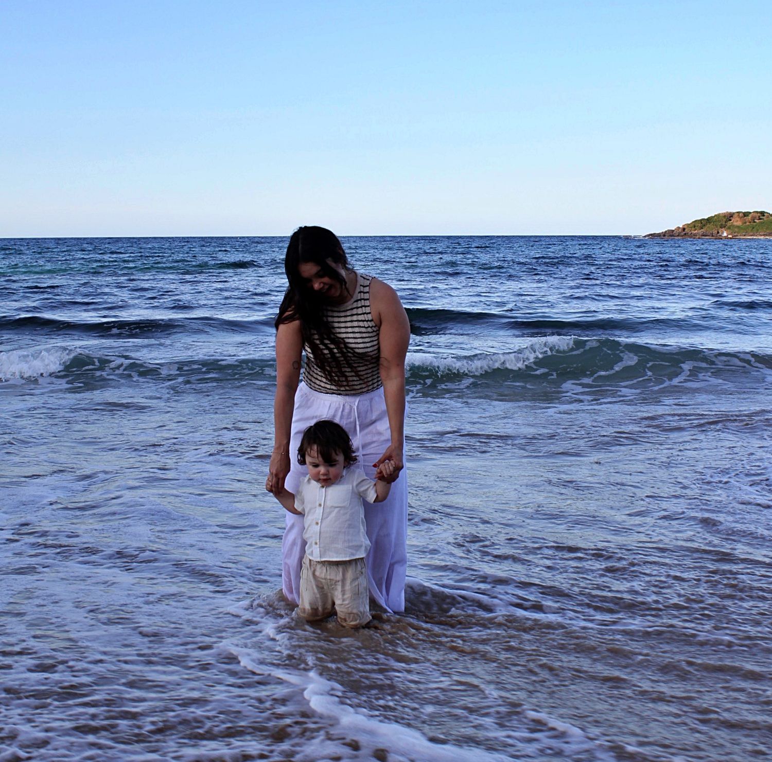 Beach motherhood session showing a mother holding her son’s hand as they play in the ocean together.