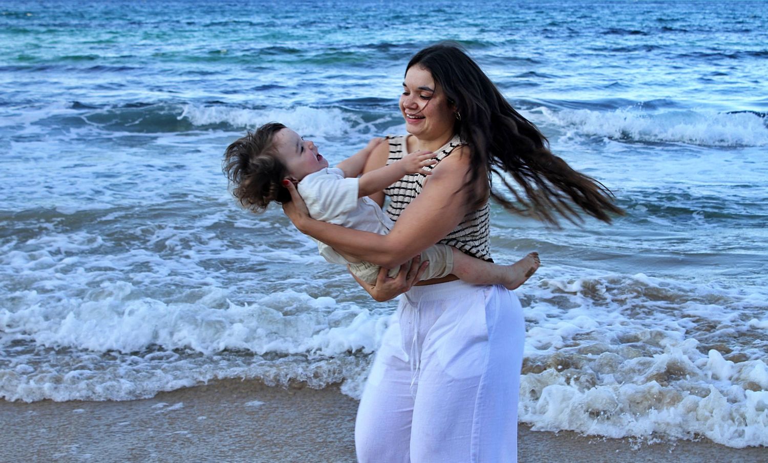 Beach motherhood session capturing a mother spinning her son while holding him, both enjoying a playful moment by the ocean.
