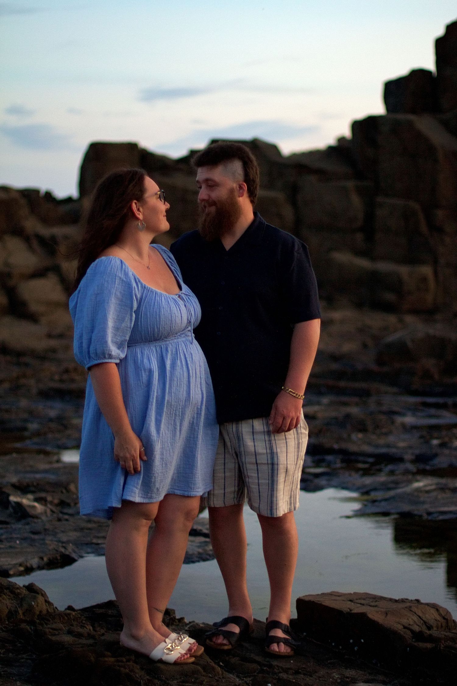 Romantic couple standing close together at Bombo Quarry, gazing at each other with dramatic rock formations and ocean behind them.