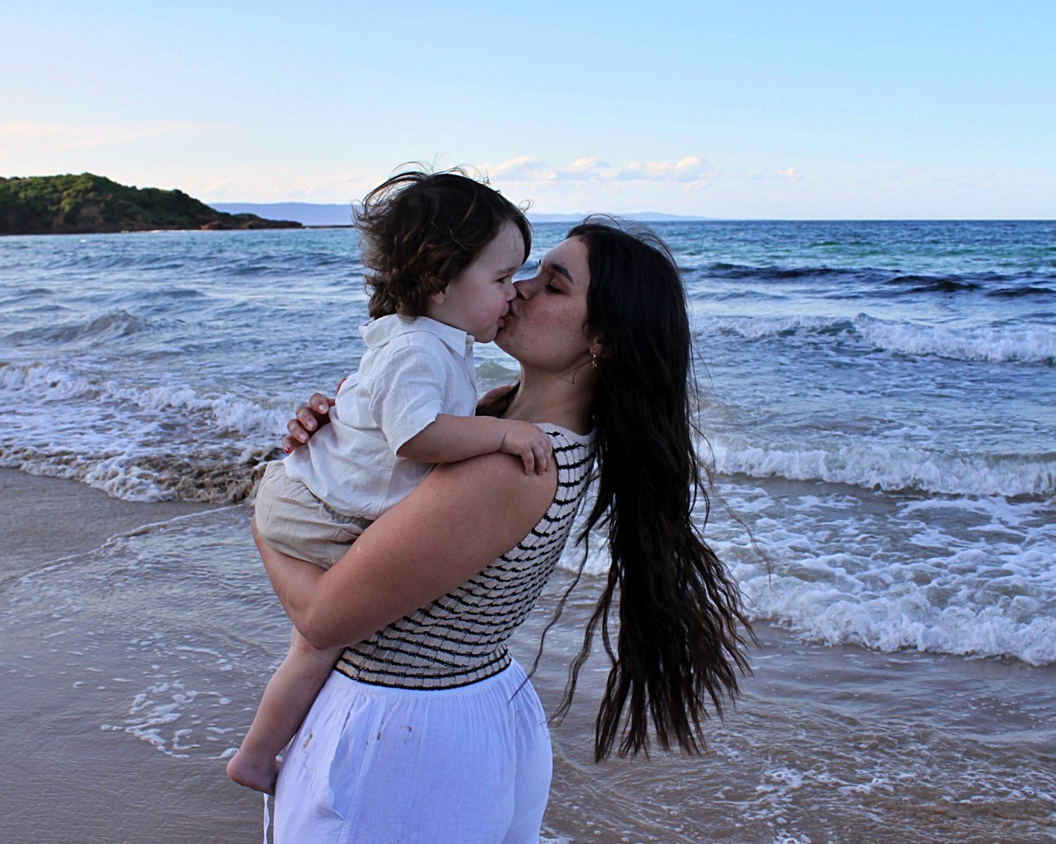 Mother holding her son and giving him a kiss with the ocean behind them during a beach motherhood session.