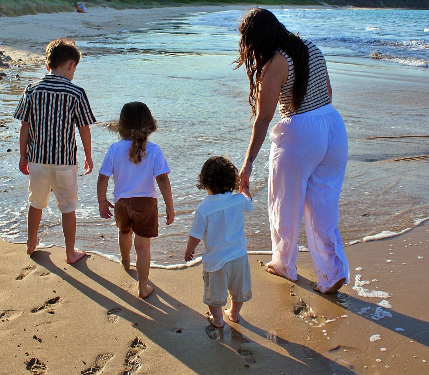 Mother walking hand in hand with her three boys along the beach, photographed from behind in natural light.