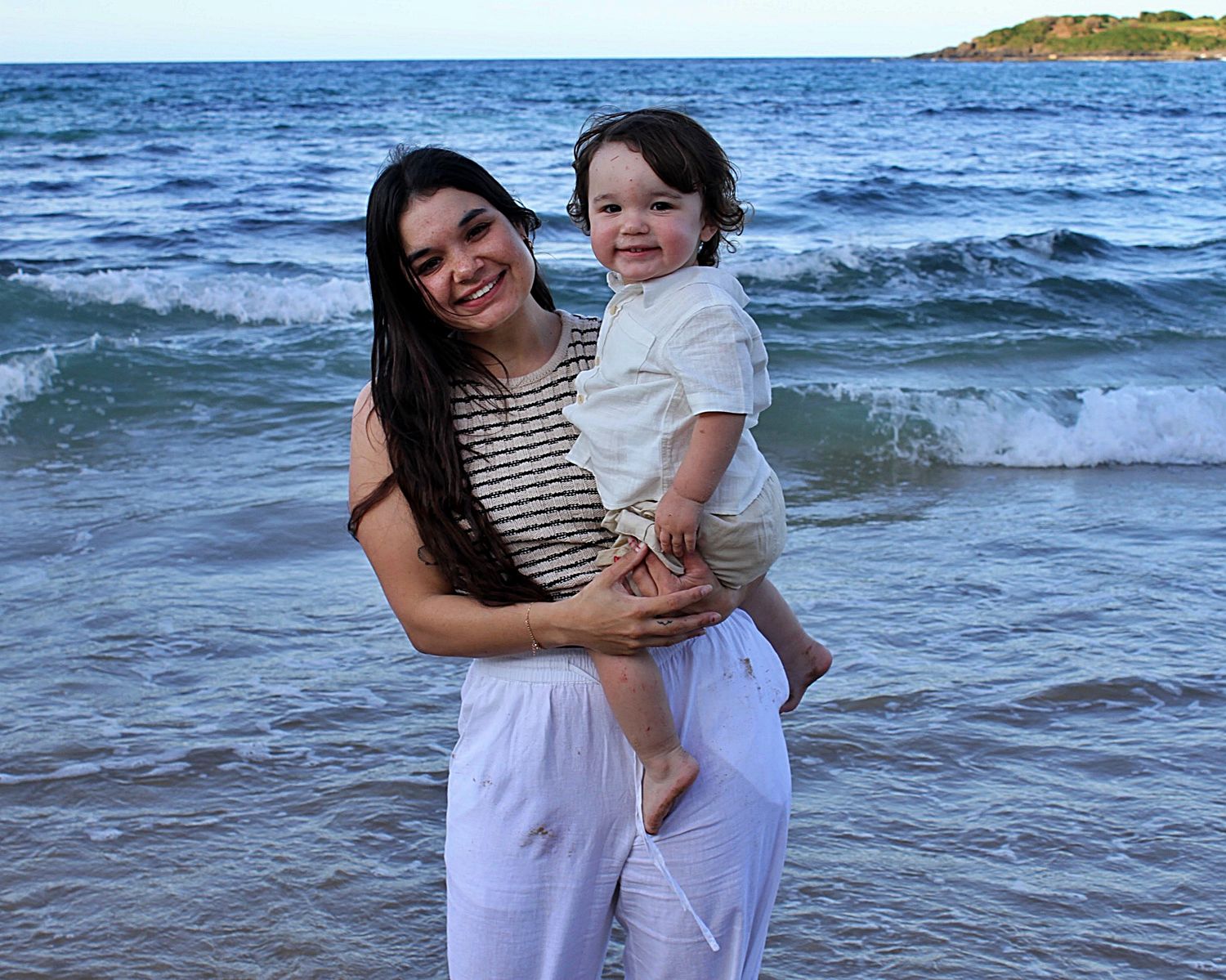 Mother holding her son, both looking at the camera smiling with the ocean behind them during a beach motherhood session.
