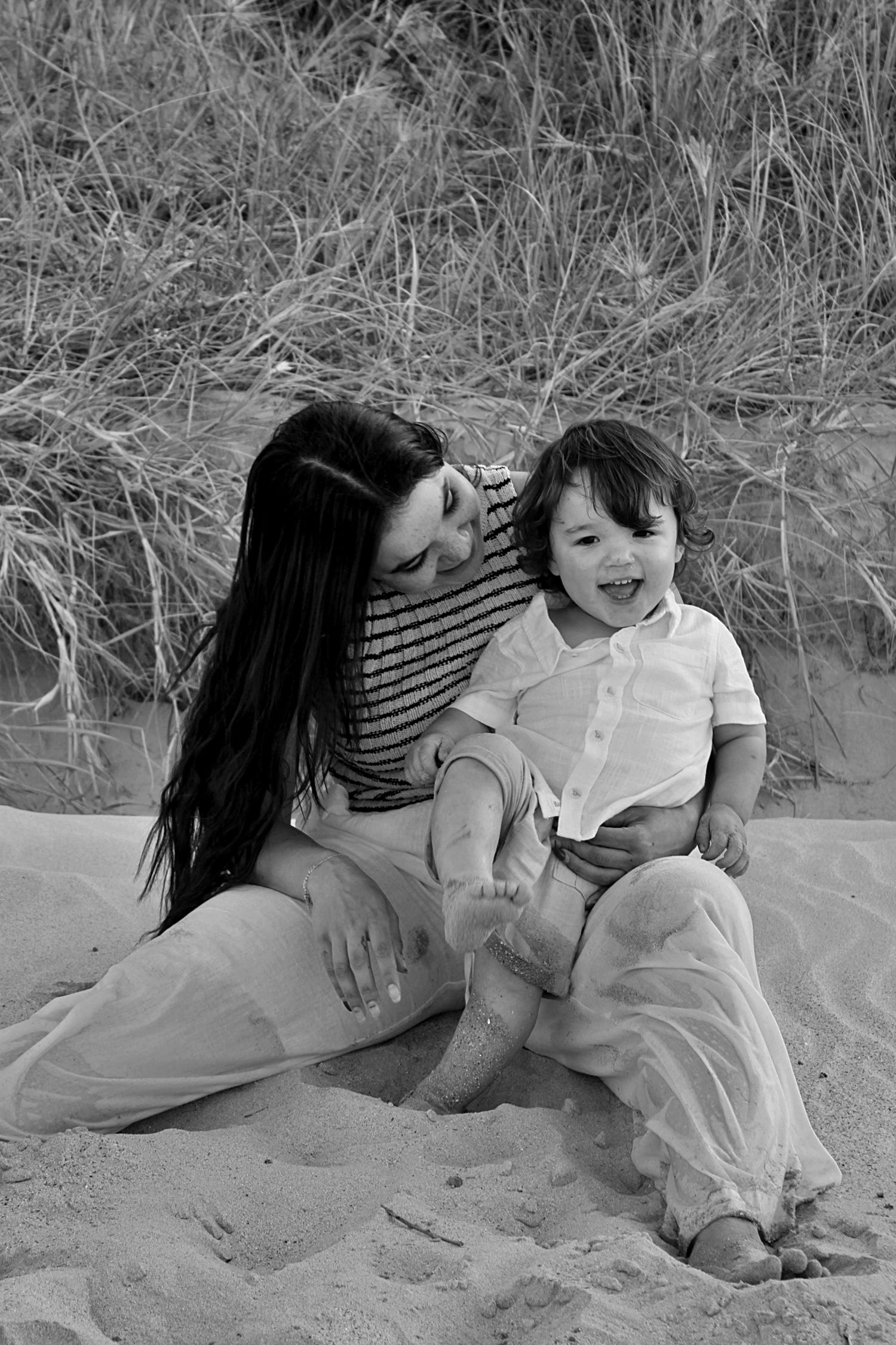 Beach motherhood session of a mother looking at her son as he smiles at the camera with grassy lands behind them