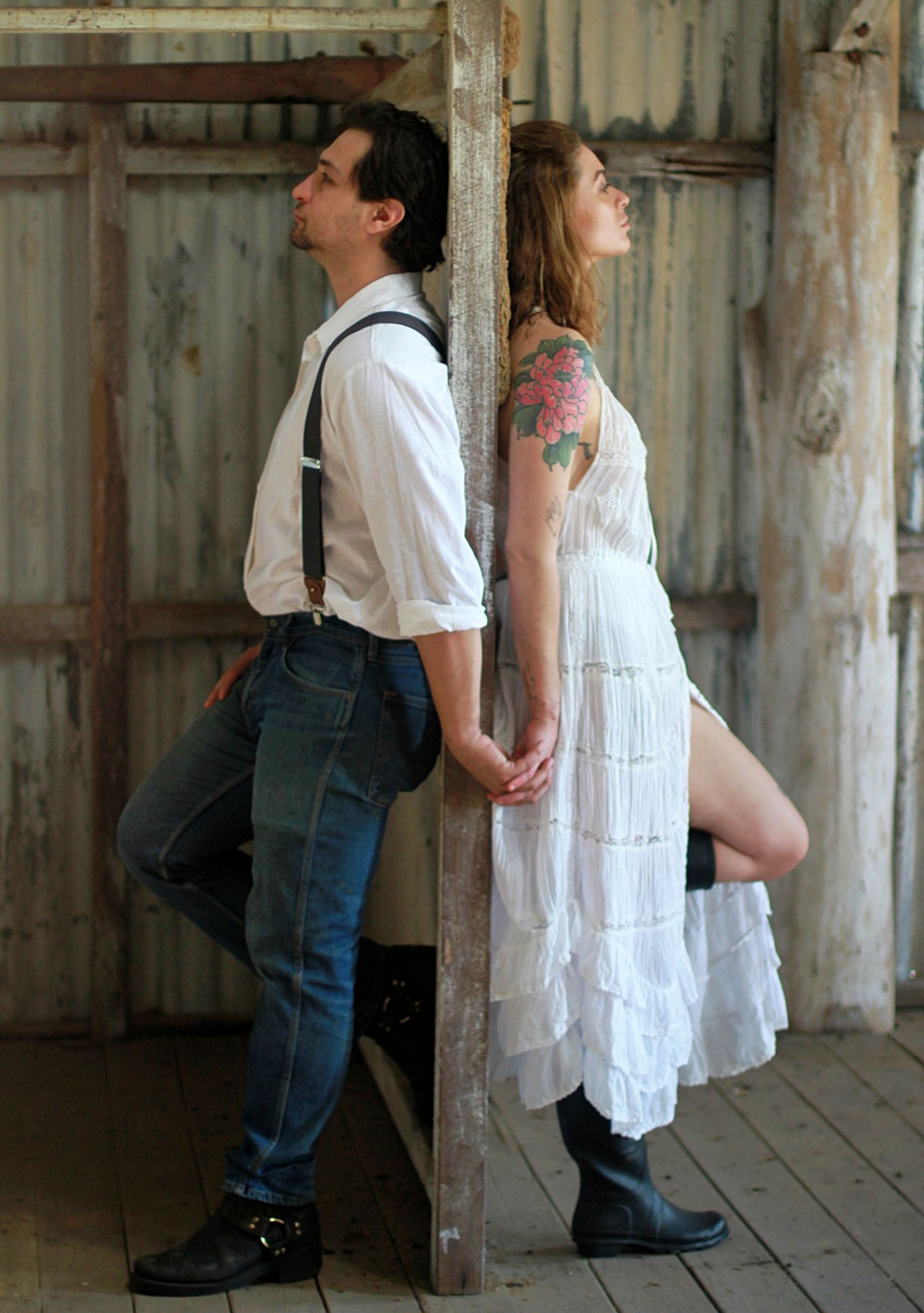 Couple holding hands through a wall, back to back, captured in a candid barn engagement photography session.