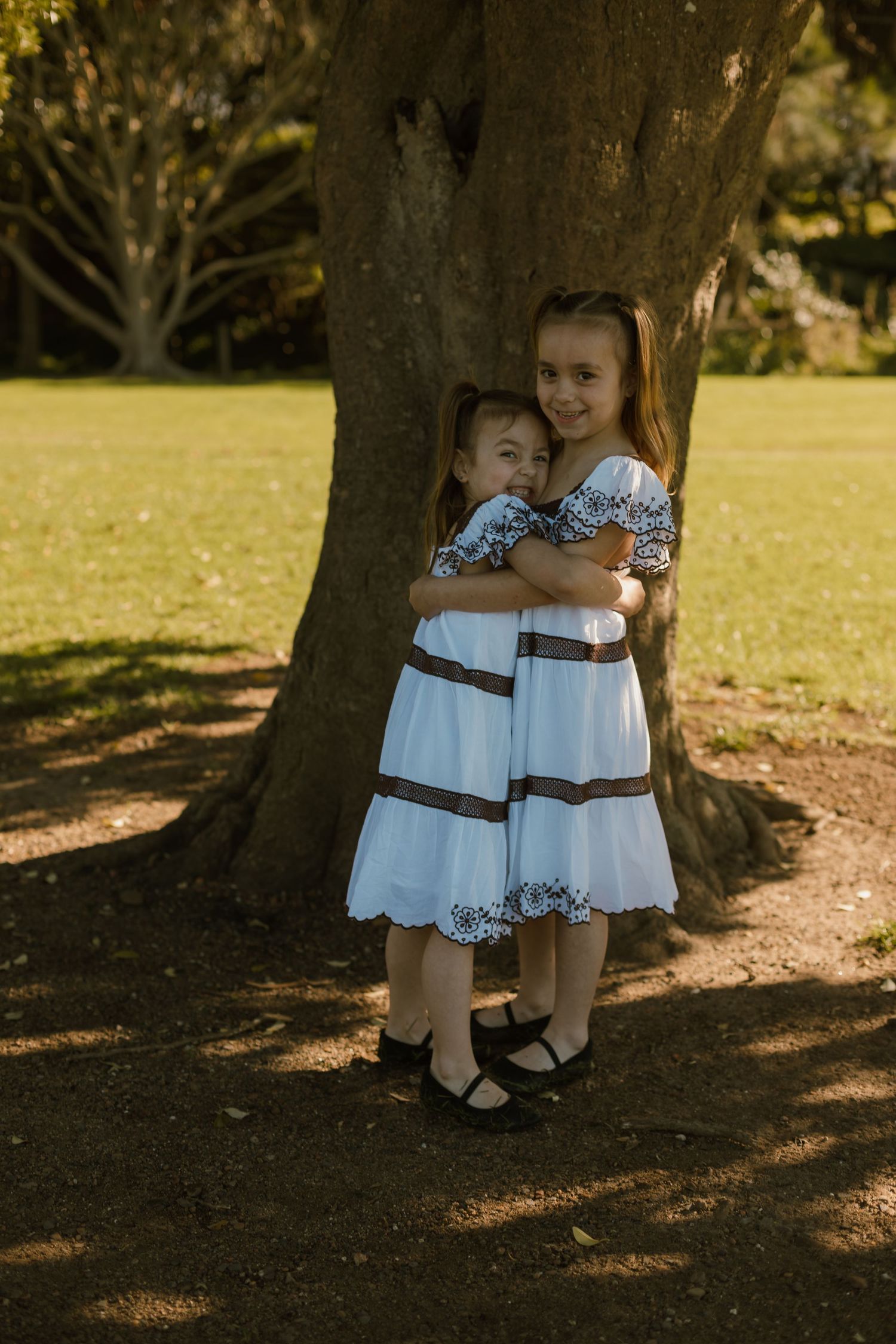Two young sisters cuddling together beneath a large tree during an outdoor family photography session.