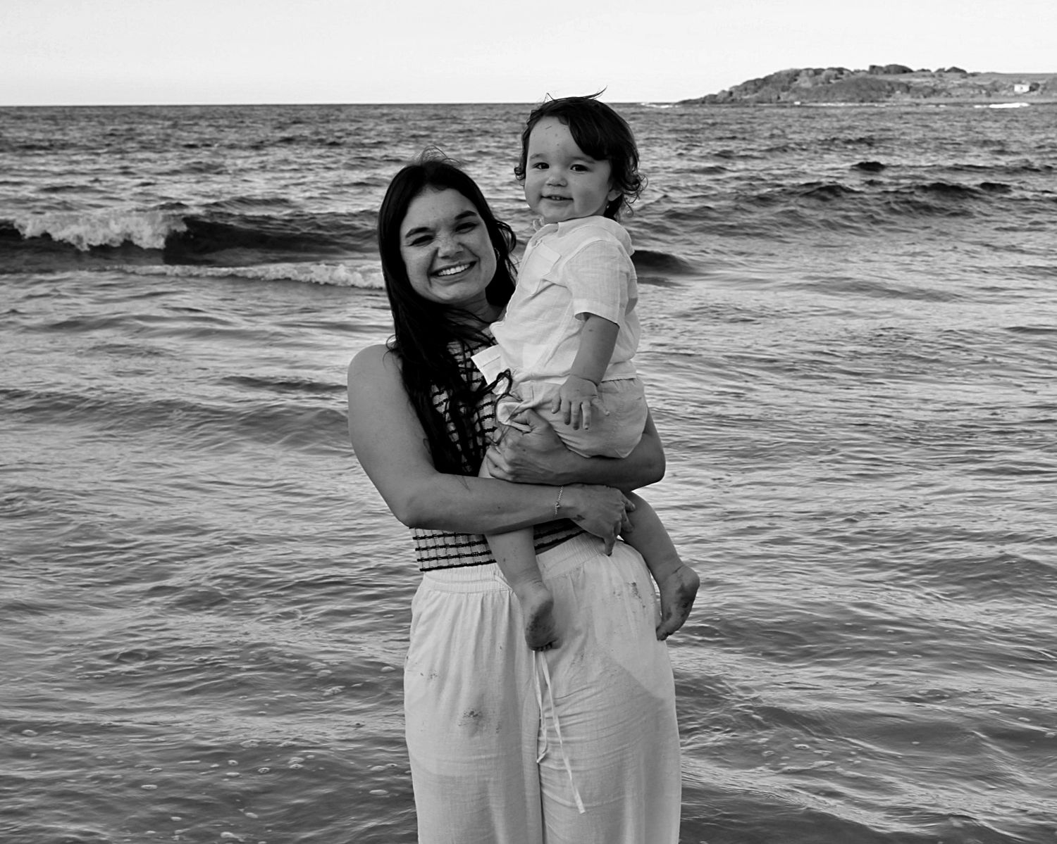 Mother holding her son, both looking at the camera smiling with the ocean behind them during a beach motherhood session.