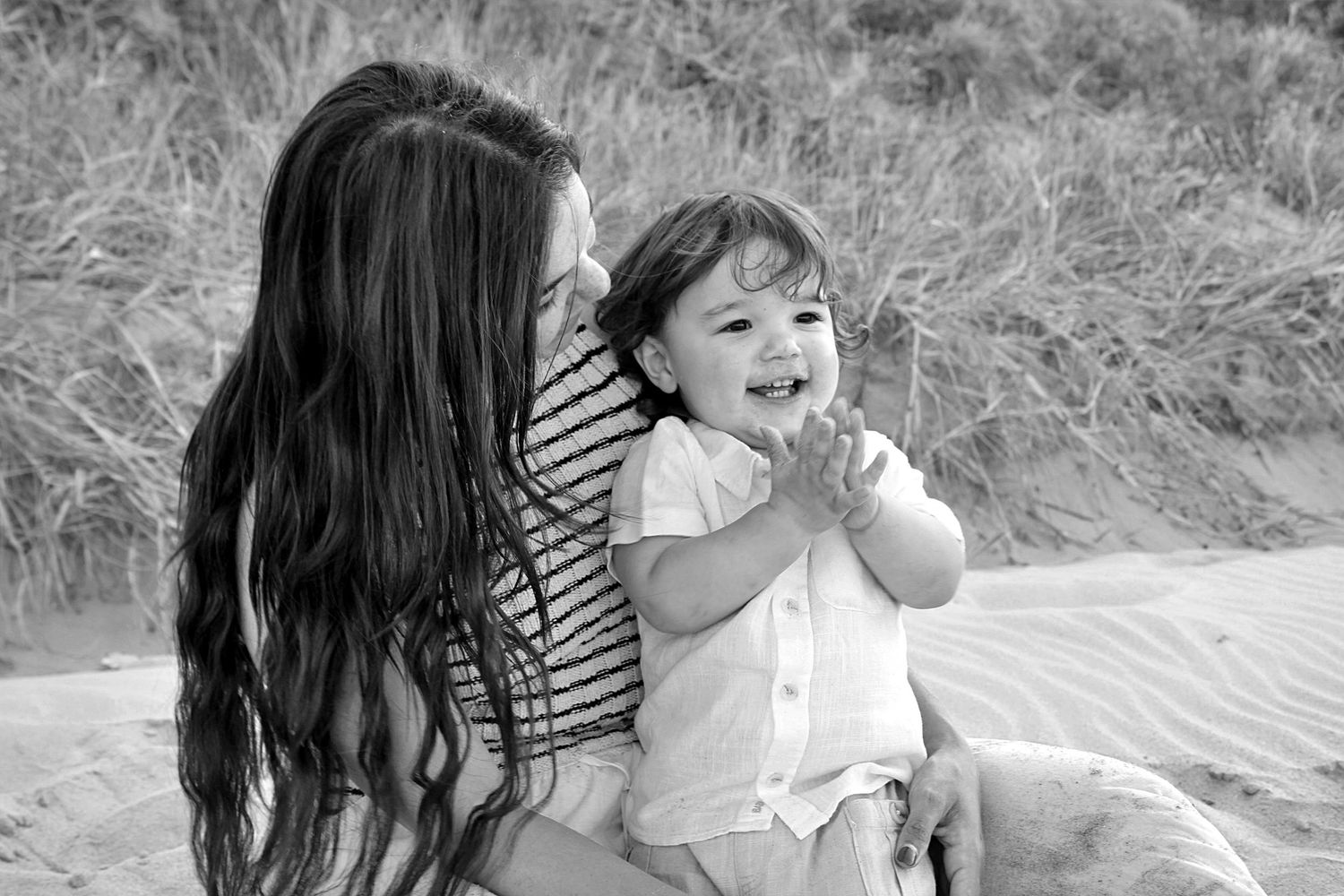 Beach motherhood session capturing a mother looking at her son while he claps excitedly on the sand.