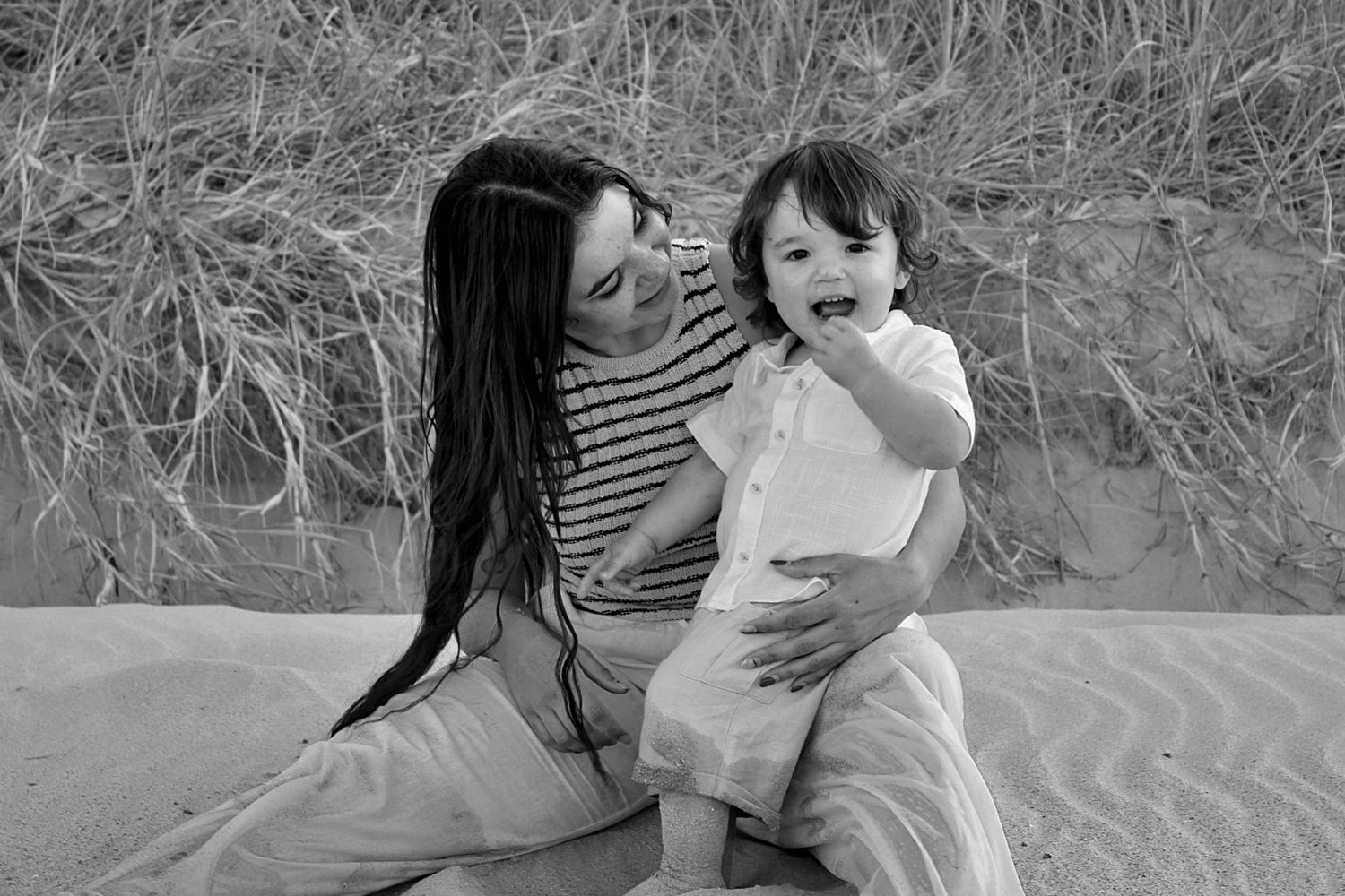 Beach motherhood session of a mother looking at her son as he smiles at the camera with grassy lands behind them.