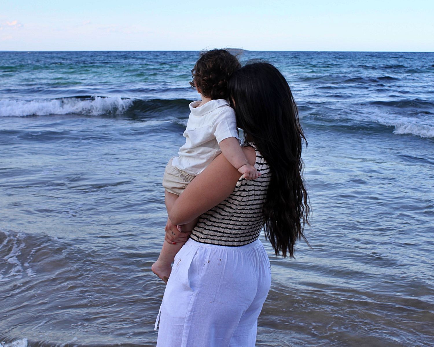 Mother holding her son on the shoreline during a beach motherhood session, looking out at the ocean, photographed from behind.