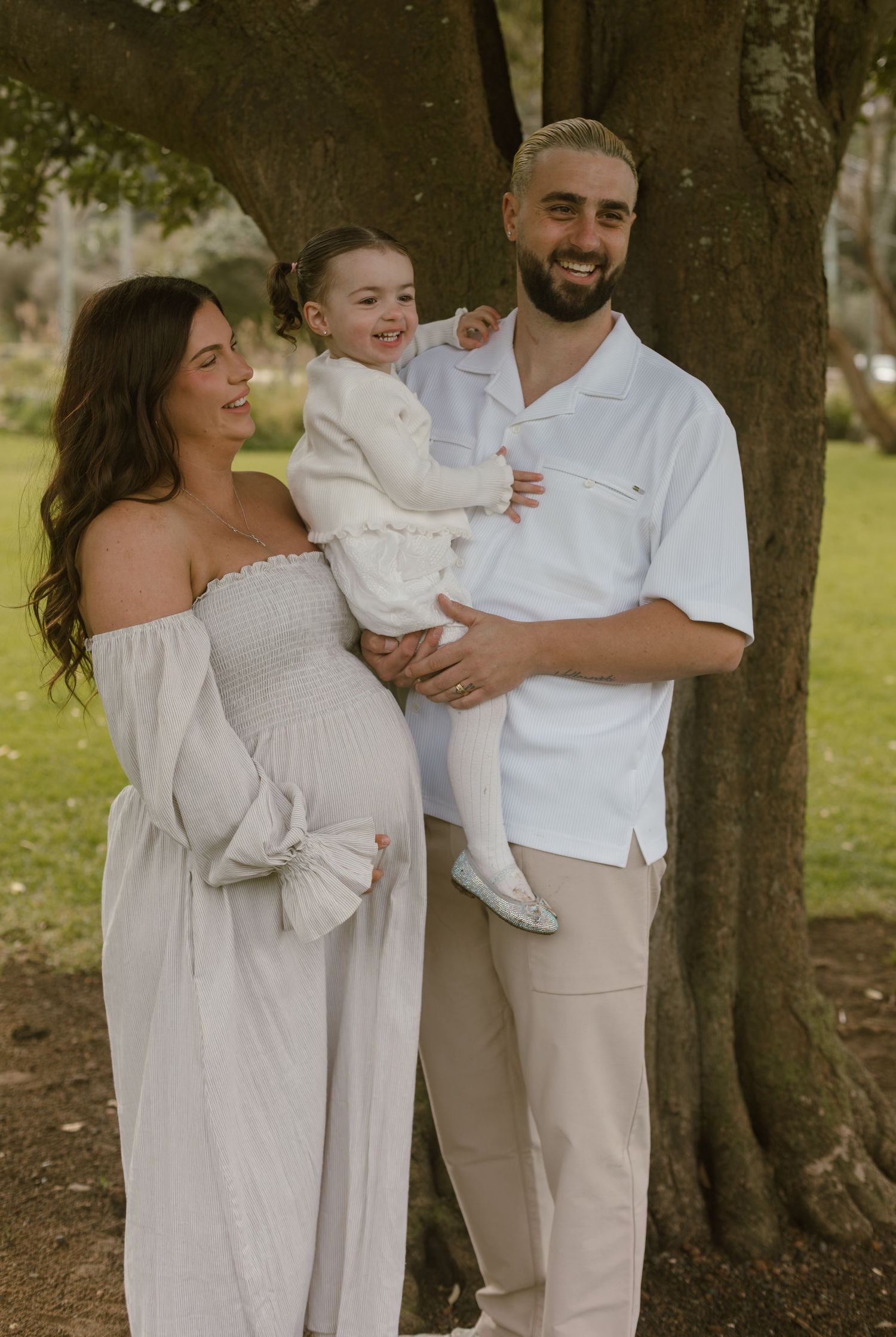 Mum and Dad holding their young daughter together beside a large tree during an outdoor family maternity session.