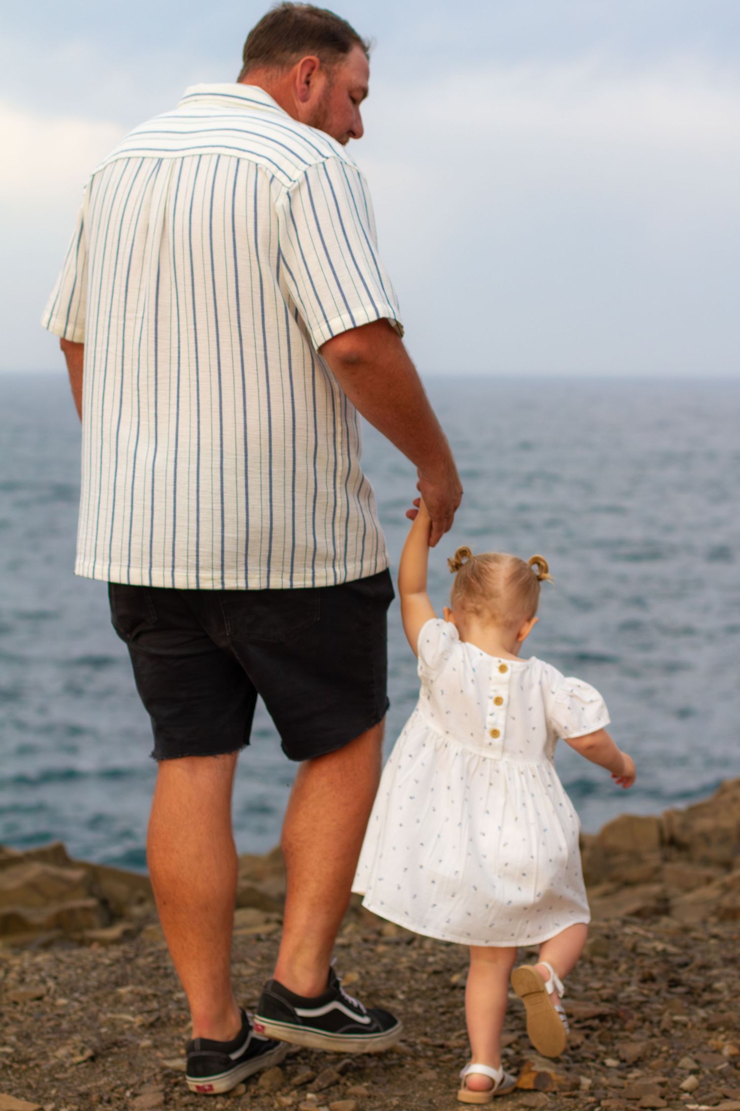 Dad holding his young daughter's hand as they walk together beside the ocean during a family photography session.