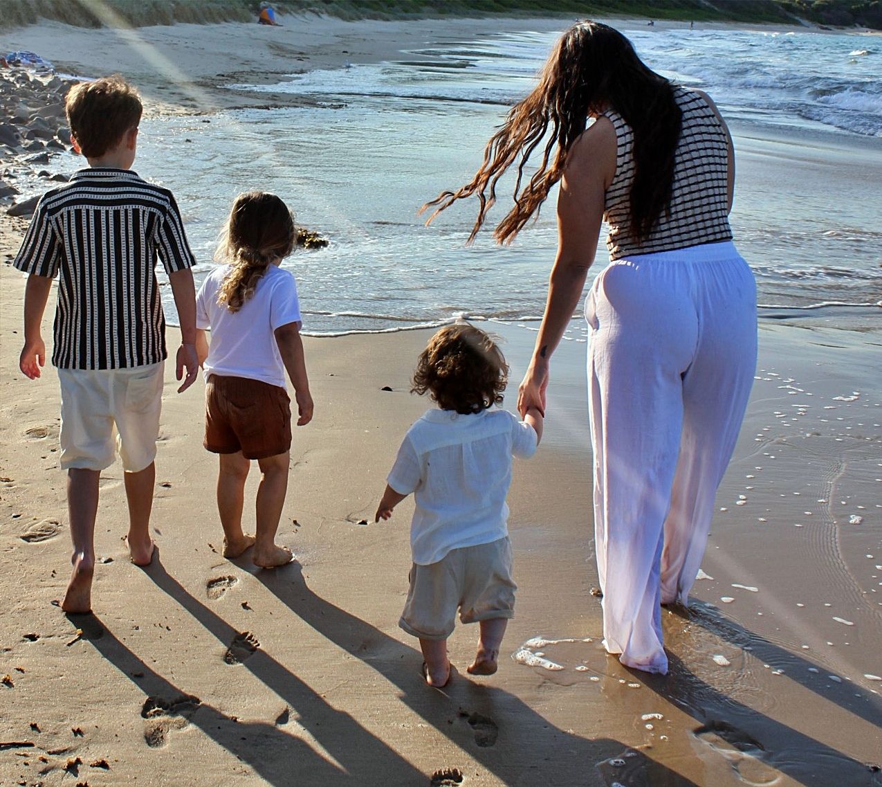 Mother walking hand in hand with her three boys along the beach, photographed from behind in natural light.
