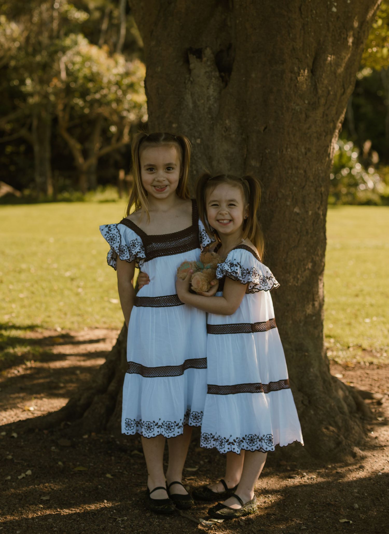 Two sisters standing together and cuddling beneath a large tree during their outdoor family session.