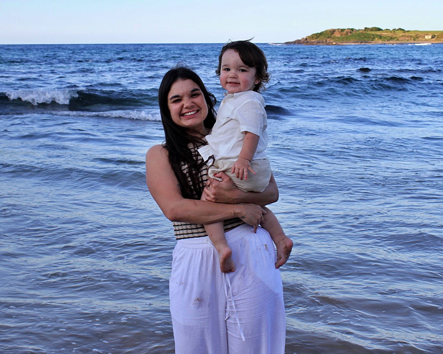 Mother holding her son, both looking at the camera smiling with the ocean behind them during a beach motherhood session.