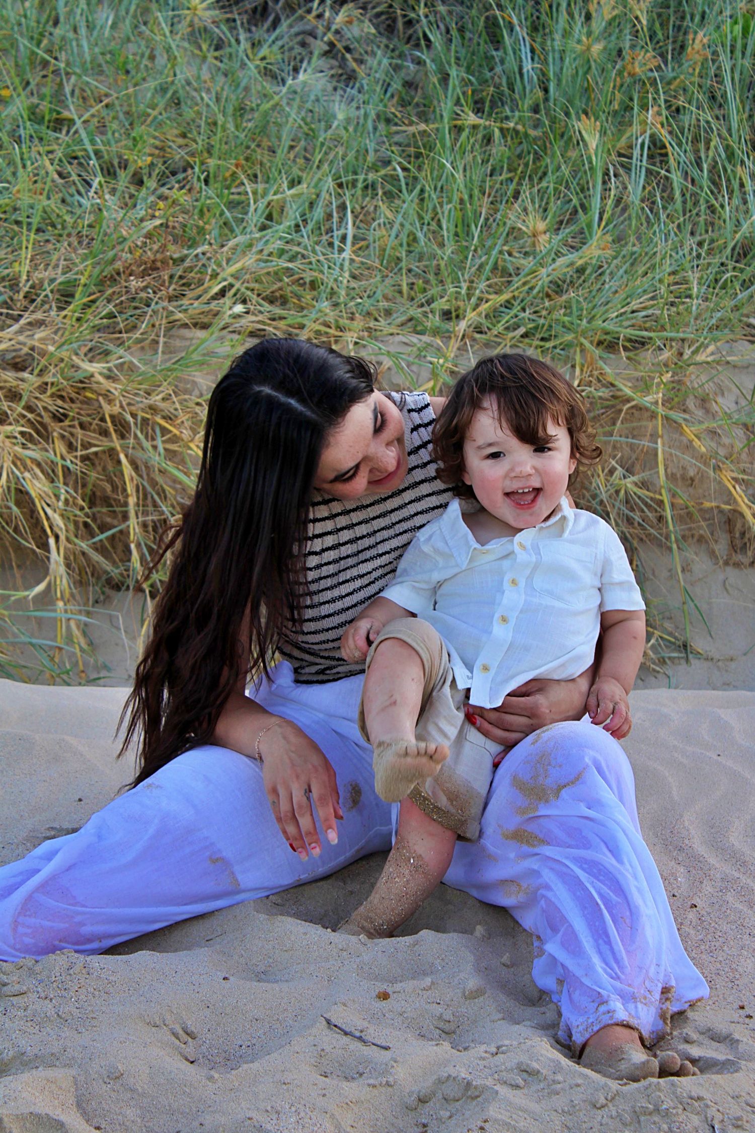 Beach motherhood session of a mother looking at her son as he smiles at the camera with grassy lands behind them