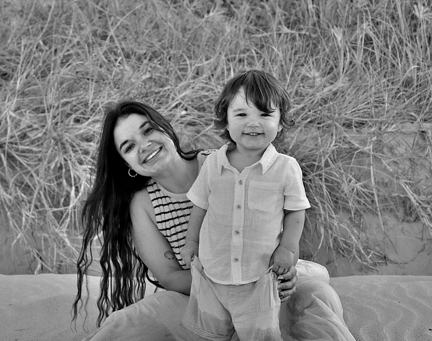 Mother and son smiling at the camera together while sitting on the beach during a motherhood session.
