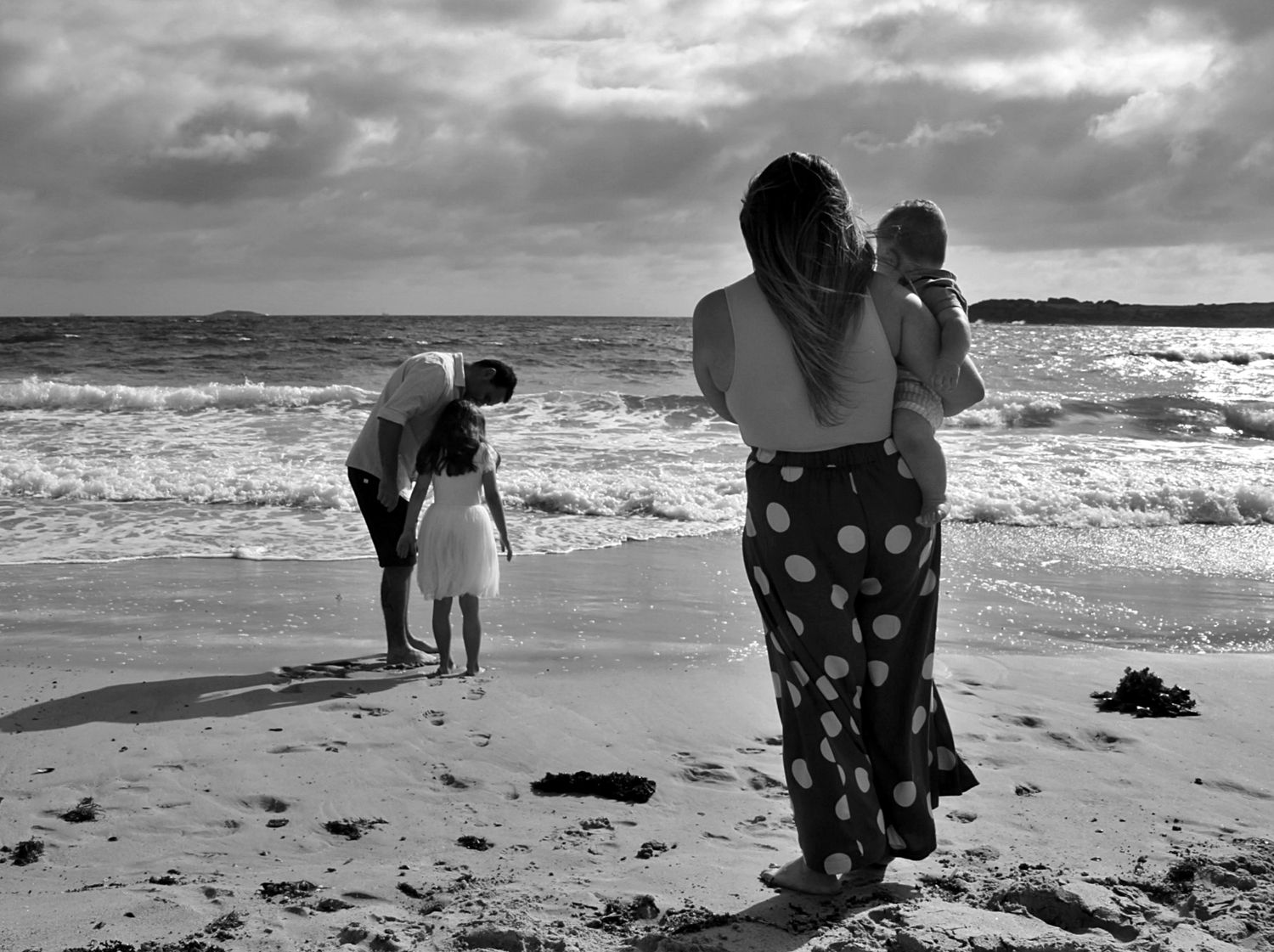 Mum and son watch dad and daughter play together on the beach in the Illawarra, capturing relaxed family connection by the sea.